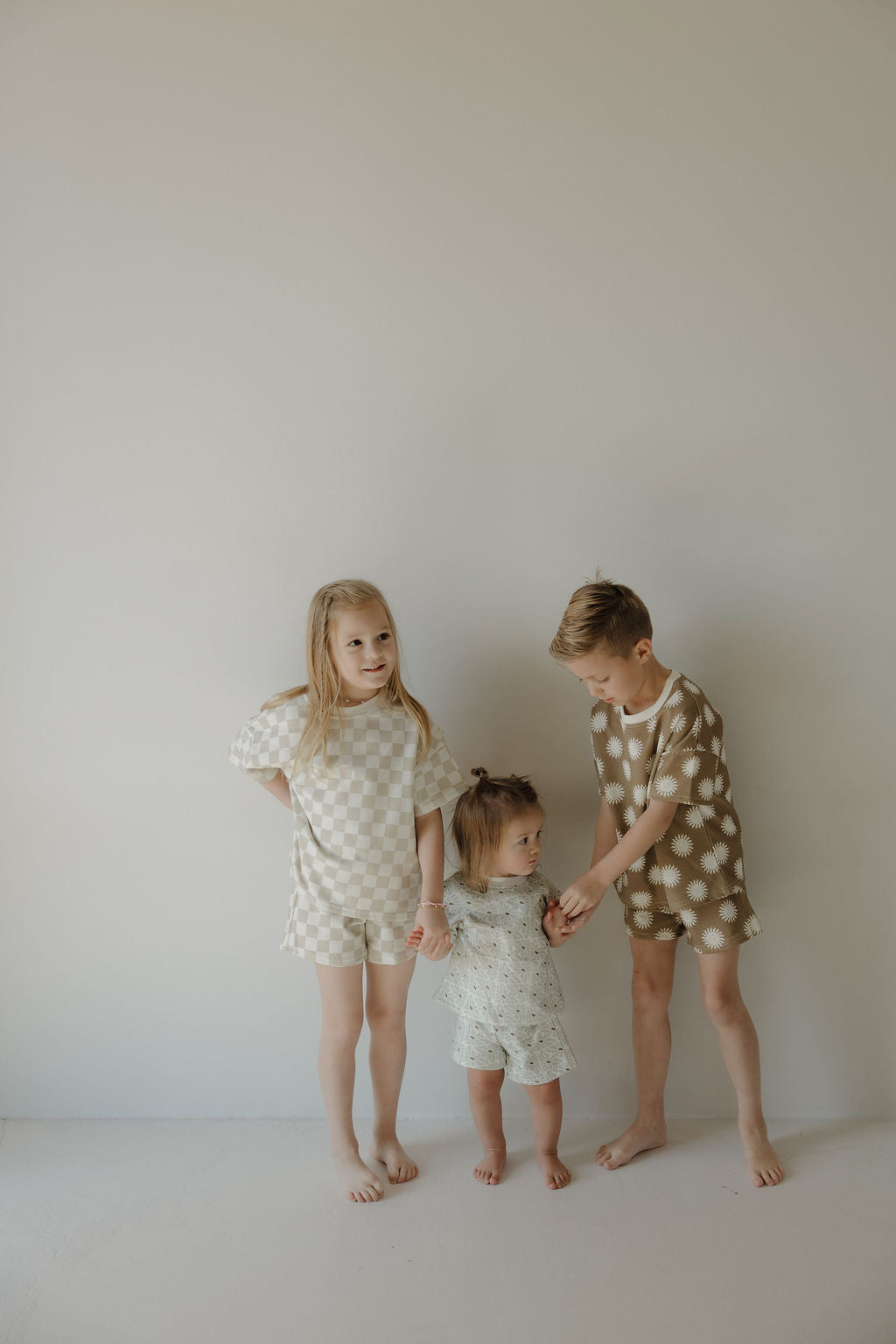 Three young children stand barefoot by a plain wall. The two older kids wear matching forever french baby French Terry Short Set | Coast outfits, while the toddler in the center wears a light ensemble. They gently hold hands and smile.