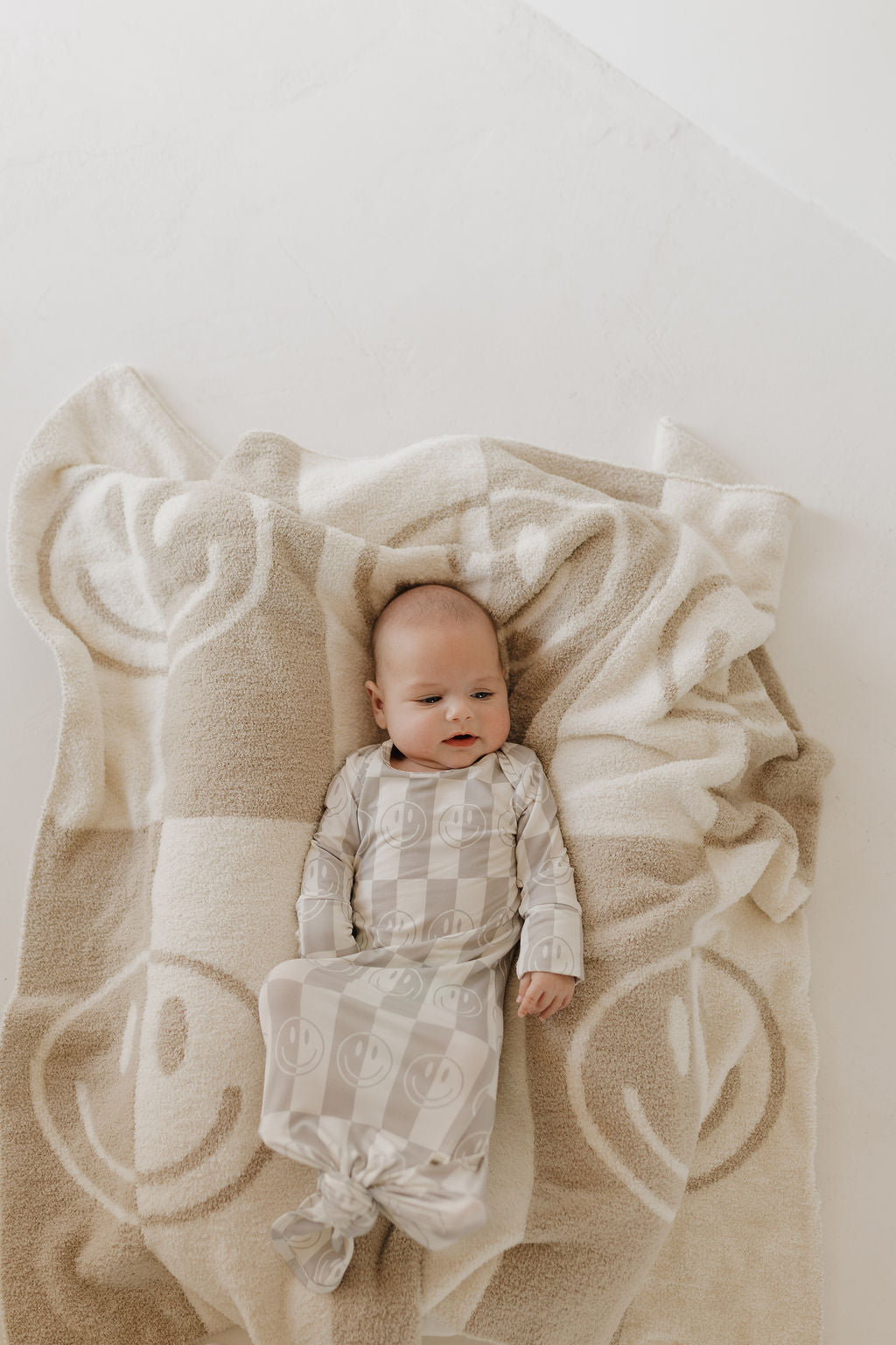 A baby is peacefully resting on a beige and white blanket, wearing the Bamboo Knotted Gown in Smile Checkerboard by forever french baby. Made from hypo-allergenic materials, this charming ensemble offers comfort on the light-colored surface.