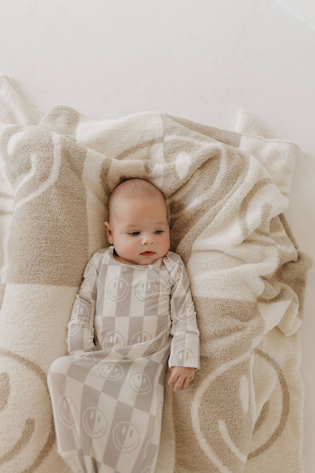 A baby lies on a soft, hypoallergenic beige and white blanket with a smiley face pattern, dressed in forever french baby's Bamboo Knotted Gown | Smile Checkerboard. The baby gazes upward with a calm expression.