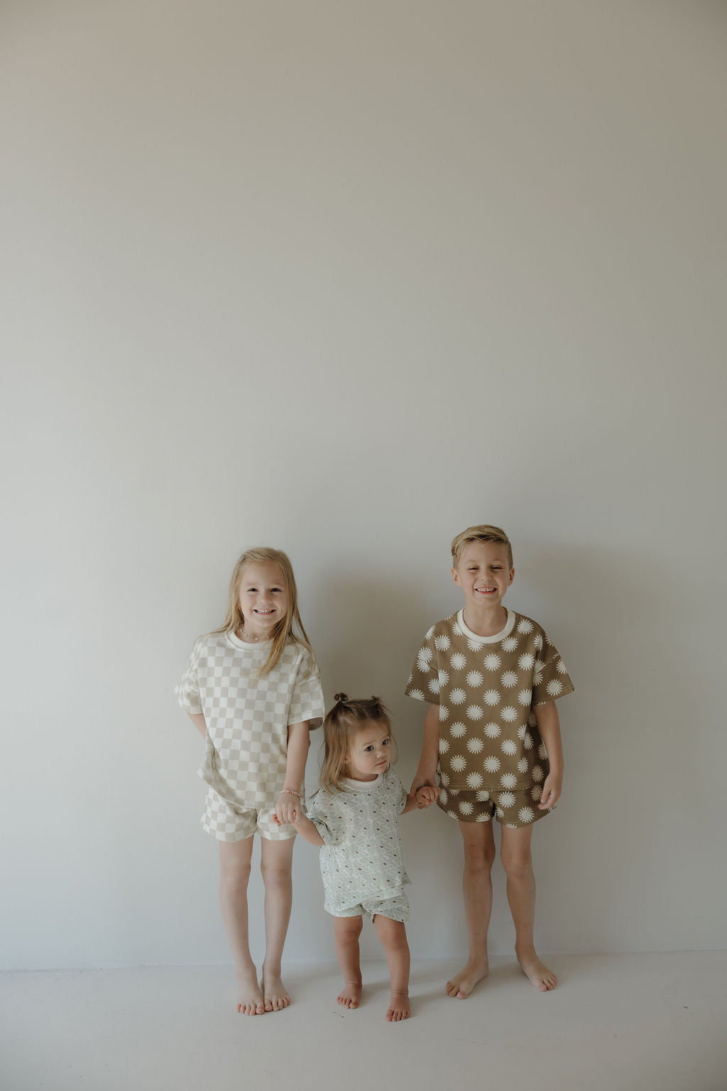 Three young children stand barefoot against a plain light-colored wall, holding hands and smiling. The two older kids wear matching forever french baby French Terry Short Sets in checkerboard, while the youngest wears a light-colored patterned outfit.