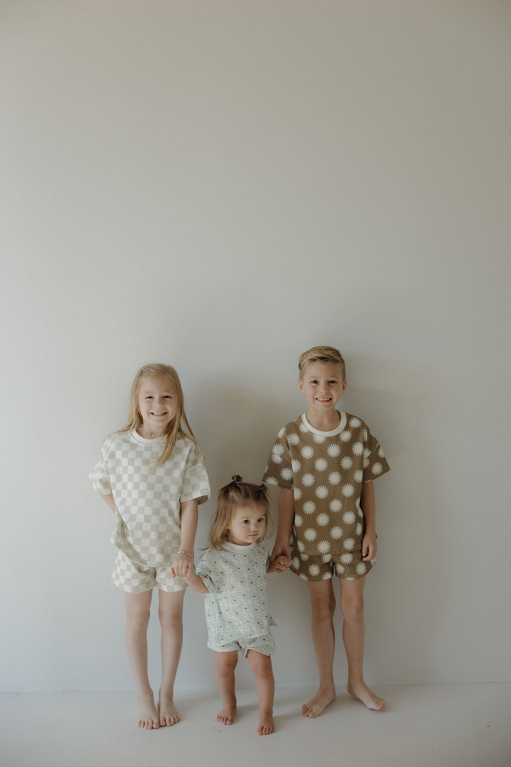 Three young children stand barefoot against a plain light wall, holding hands. The two older kids wear matching forever french baby French Terry Short Set | Sol outfits, while the youngest stands between them in a light printed ensemble.