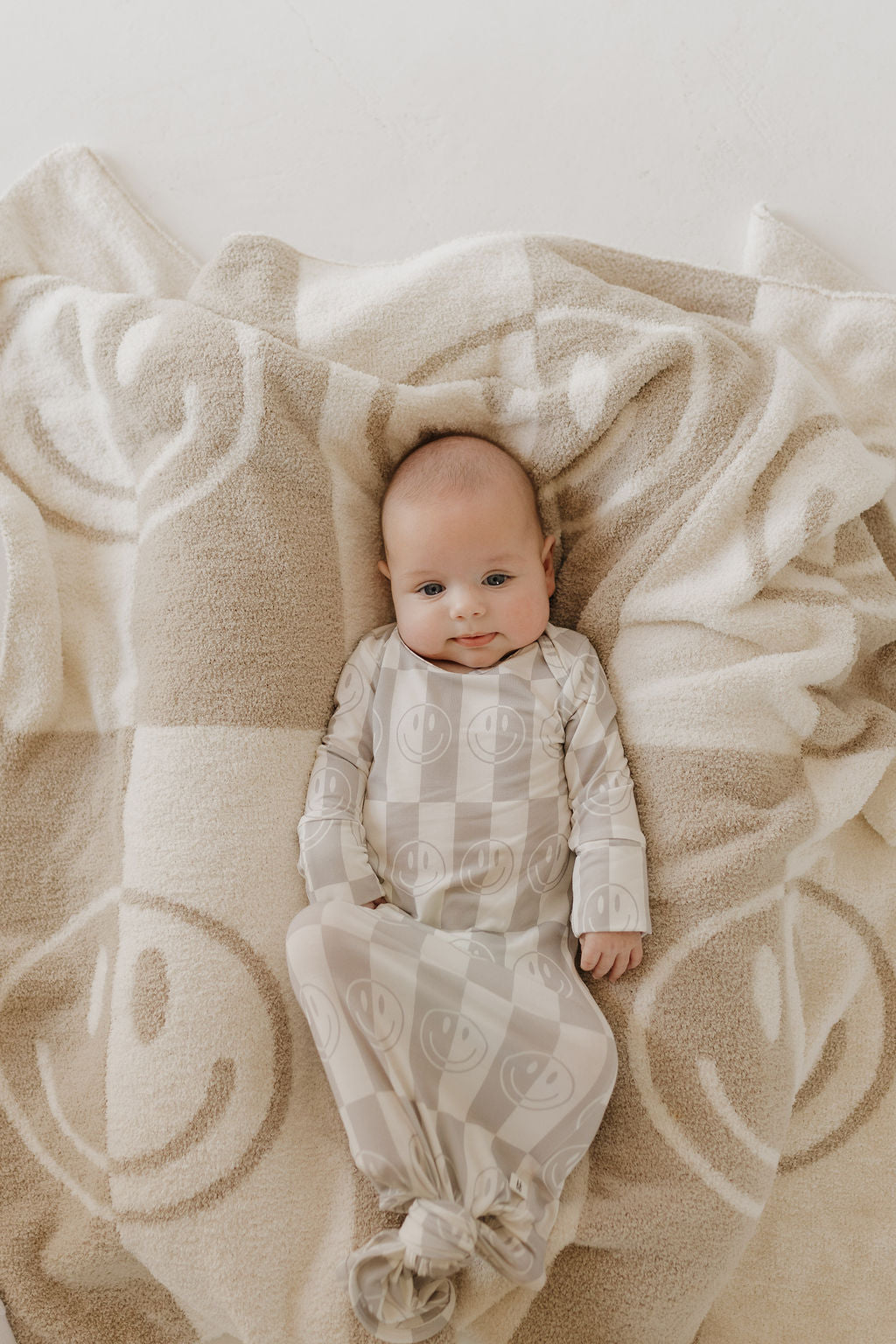 A baby lies content in a soft, hypoallergenic Bamboo Knotted Gown | Smile Checkerboard by forever french baby, enjoying the cozy comfort of this beige onesie adorned with smiley patterns.