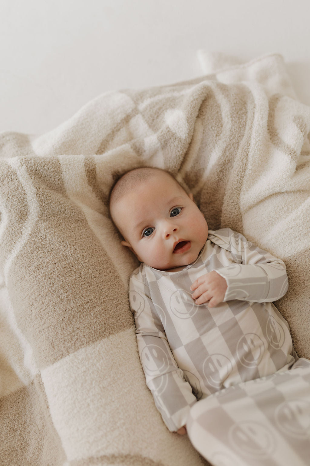 A baby lays on a beige and white blanket in a forever french baby Bamboo Knotted Gown | Smile Checkerboard, gazing upwards with serenity, enveloped in hypoallergenic fabrics and cozy textures.
