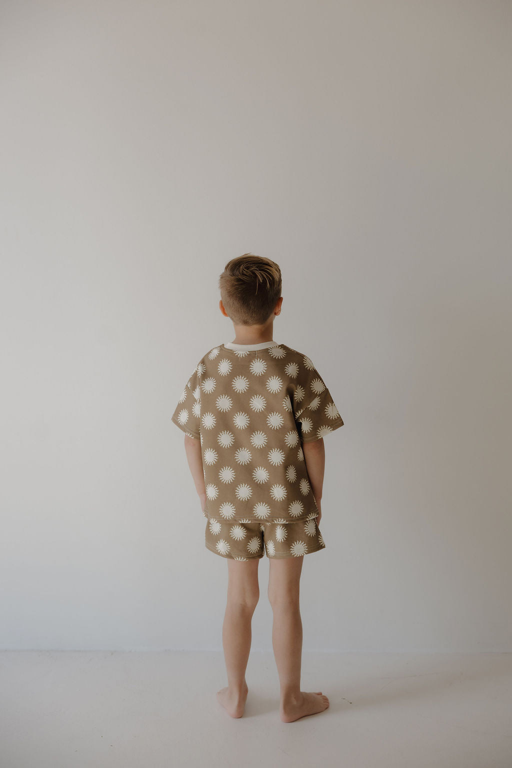 A young boy with short hair stands barefoot facing a light wall, wearing the forever french baby French Terry Short Set | Sol, crafted from a soft cotton blend with a white daisy pattern. His hands rest at his sides.