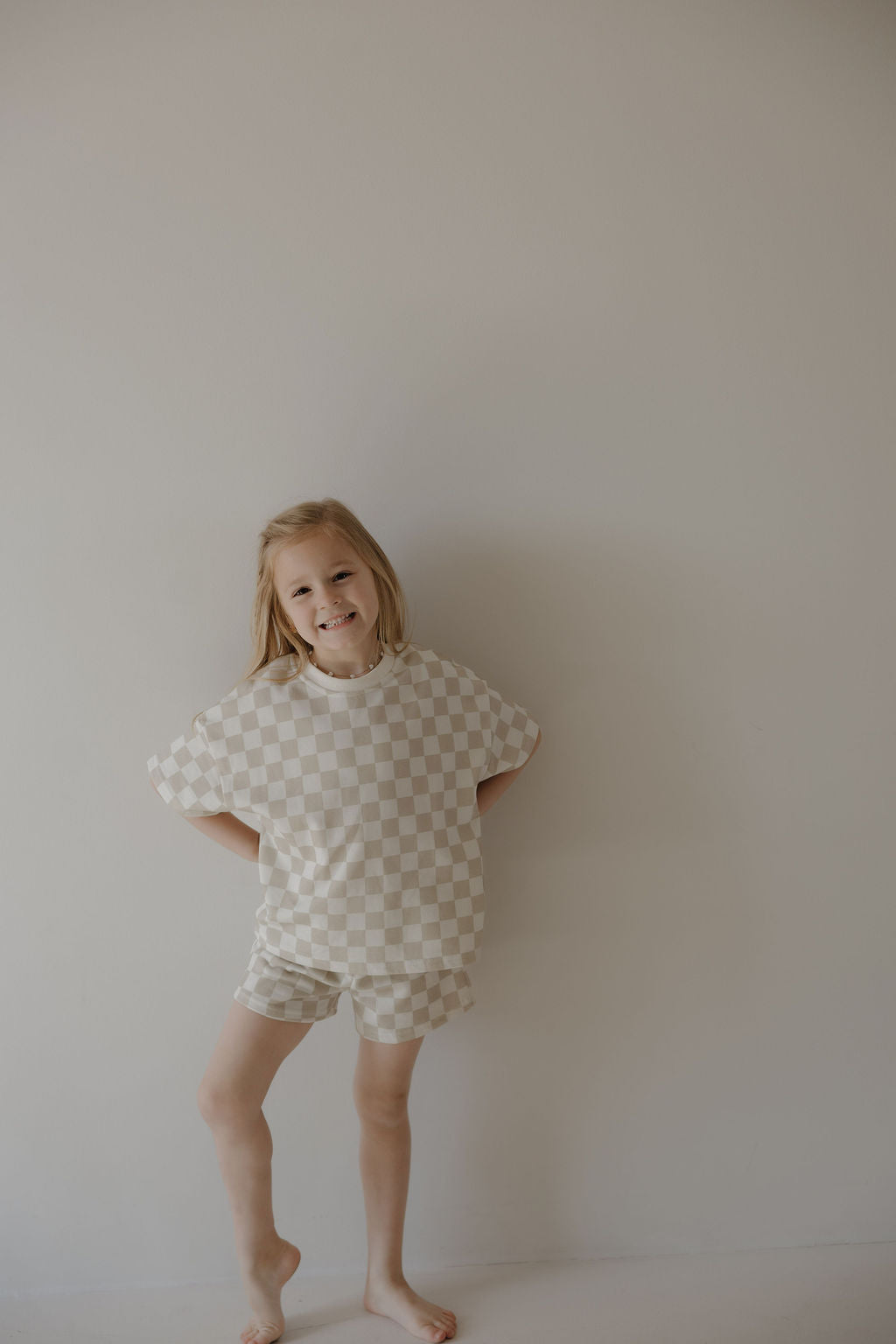A young child smiles barefoot against a light wall, wearing the forever french baby French Terry Short Set | Checkerboard—a beige and white checkered short sleeve top with matching shorts.