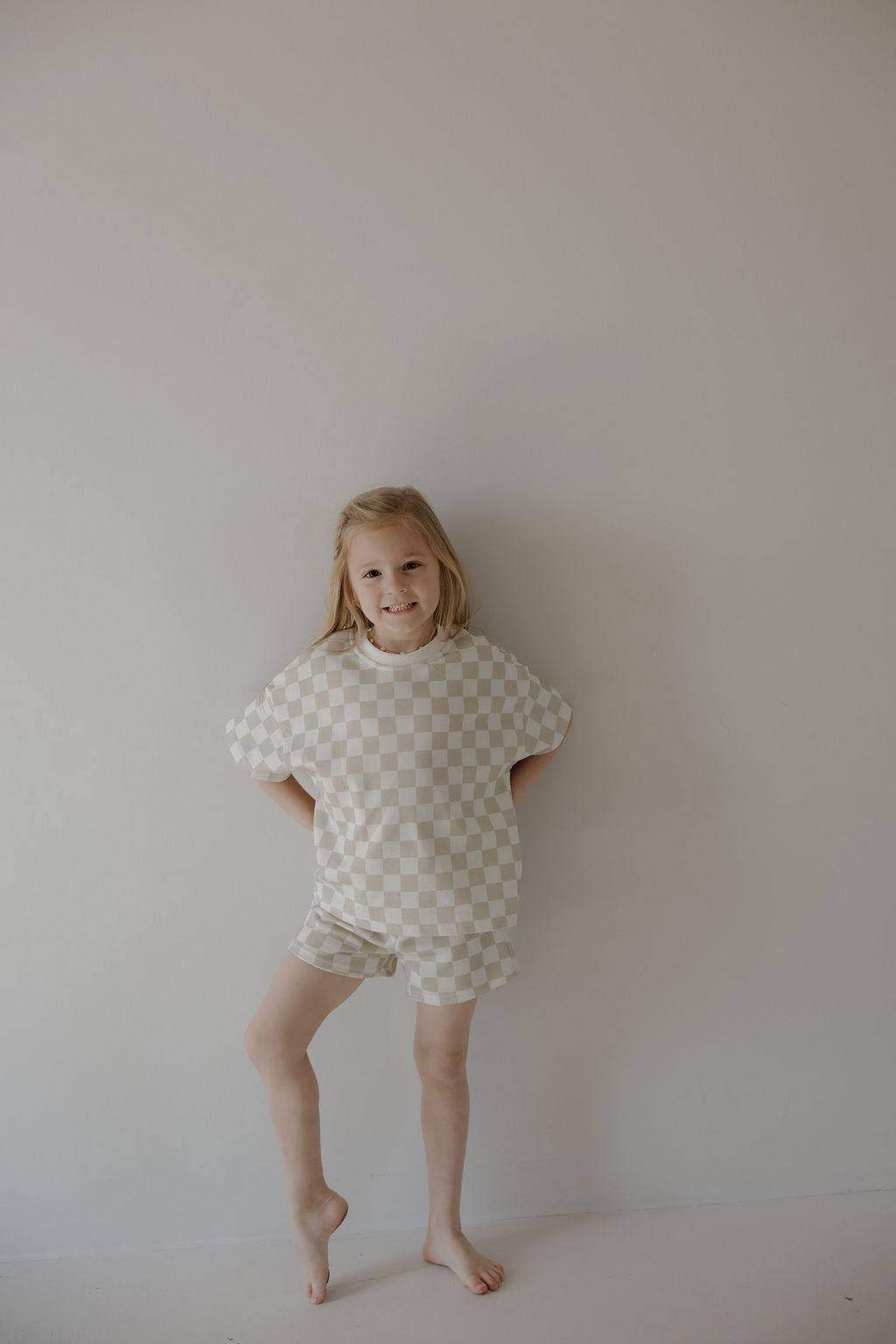 A young child with blonde hair stands barefoot against a light wall, smiling and wearing the forever french baby French Terry Short Set | Checkerboard—a soft beige and white checkered short sleeve top with matching shorts.
