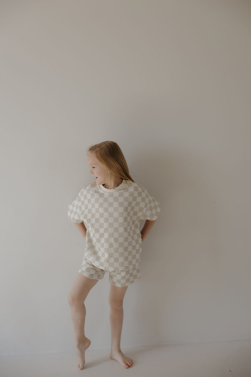 A young girl stands barefoot by a light-colored wall, smiling to the side. She’s wearing the forever french baby French Terry Short Set | Checkerboard, a soft, short-sleeve beige and white checkered matching outfit.