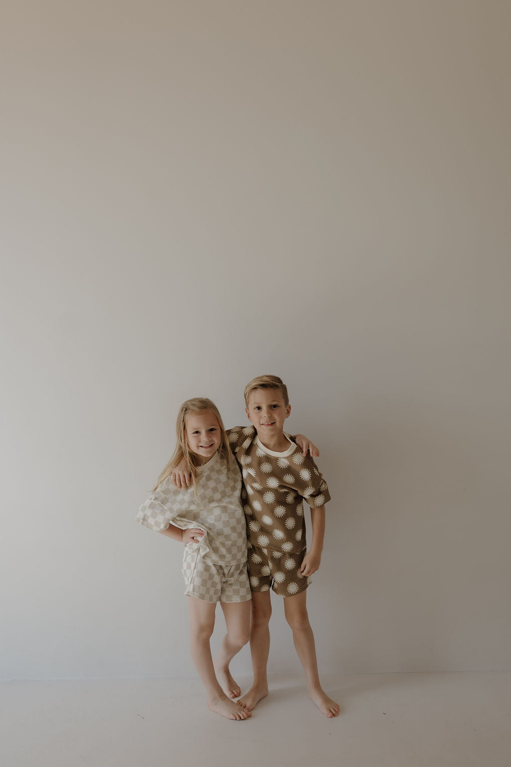Two kids smile barefoot against a plain wall in matching forever french baby pajamas. The boy wears the French Terry Short Set | Sol, a brown FF logo top and shorts, with his arm around the girl in light beige pajamas.