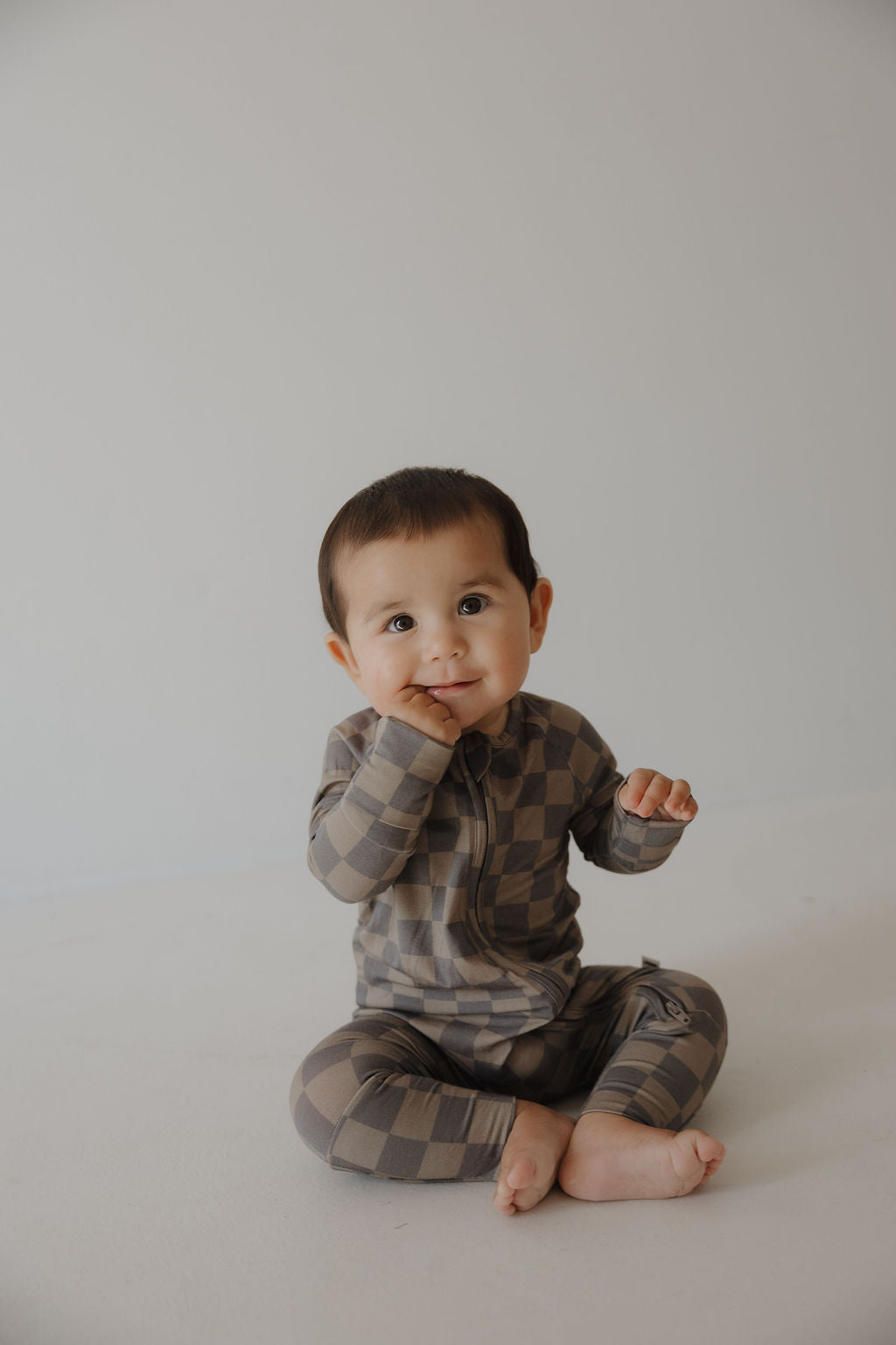 A baby with short dark hair sits on the floor, touching their mouth and looking up curiously, dressed in forever french baby's Bamboo Zip Pajamas in Faded Brown Checkerboard.