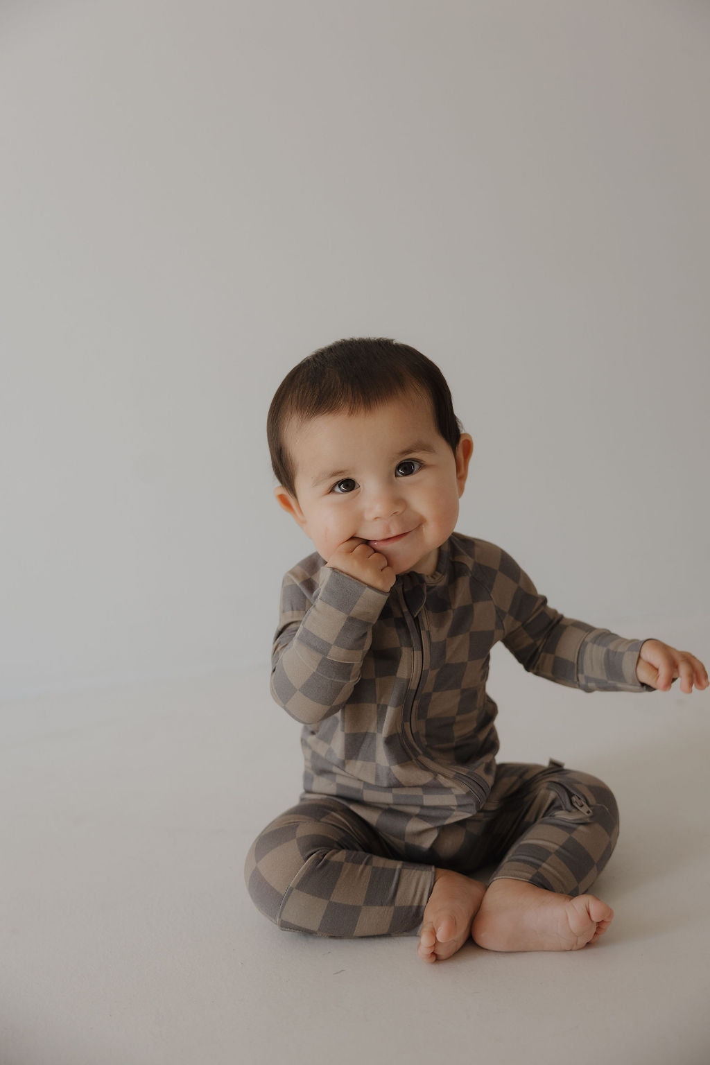 A smiling baby in forever french baby's Bamboo Zip Pajamas in Faded Brown Checkerboard, made from breathable fabric, sits on the floor with one hand on their cheek against a plain light background.