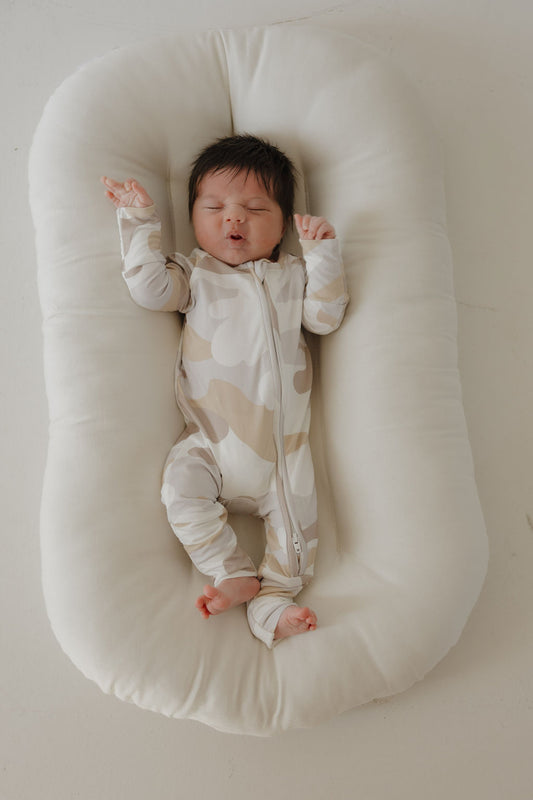 A baby with dark hair lies peacefully on a soft white cushion, eyes closed and looking relaxed, wearing forever french baby's Bamboo Zip Pajamas | Camo 2.0.