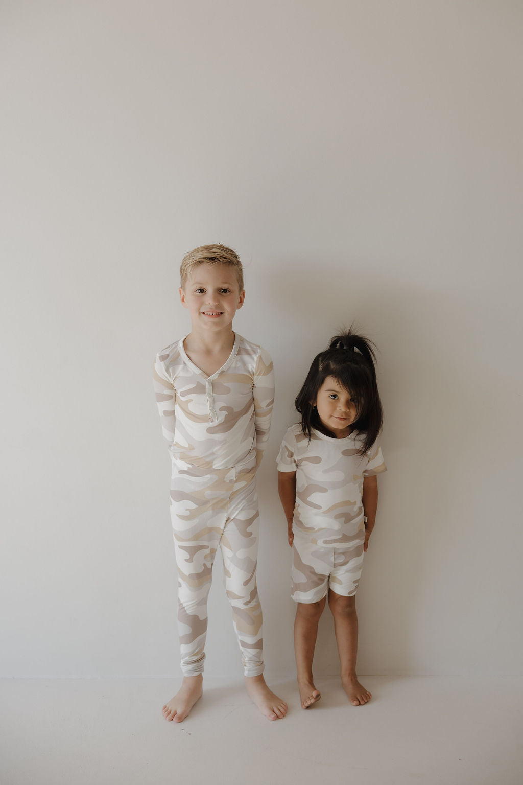 Two young children stand side by side in matching forever french baby Bamboo Two Piece Pajamas | Camo 2.0, made from hypo-allergenic bamboo for comfort—one smiles while the other has a neutral expression against a light wall.