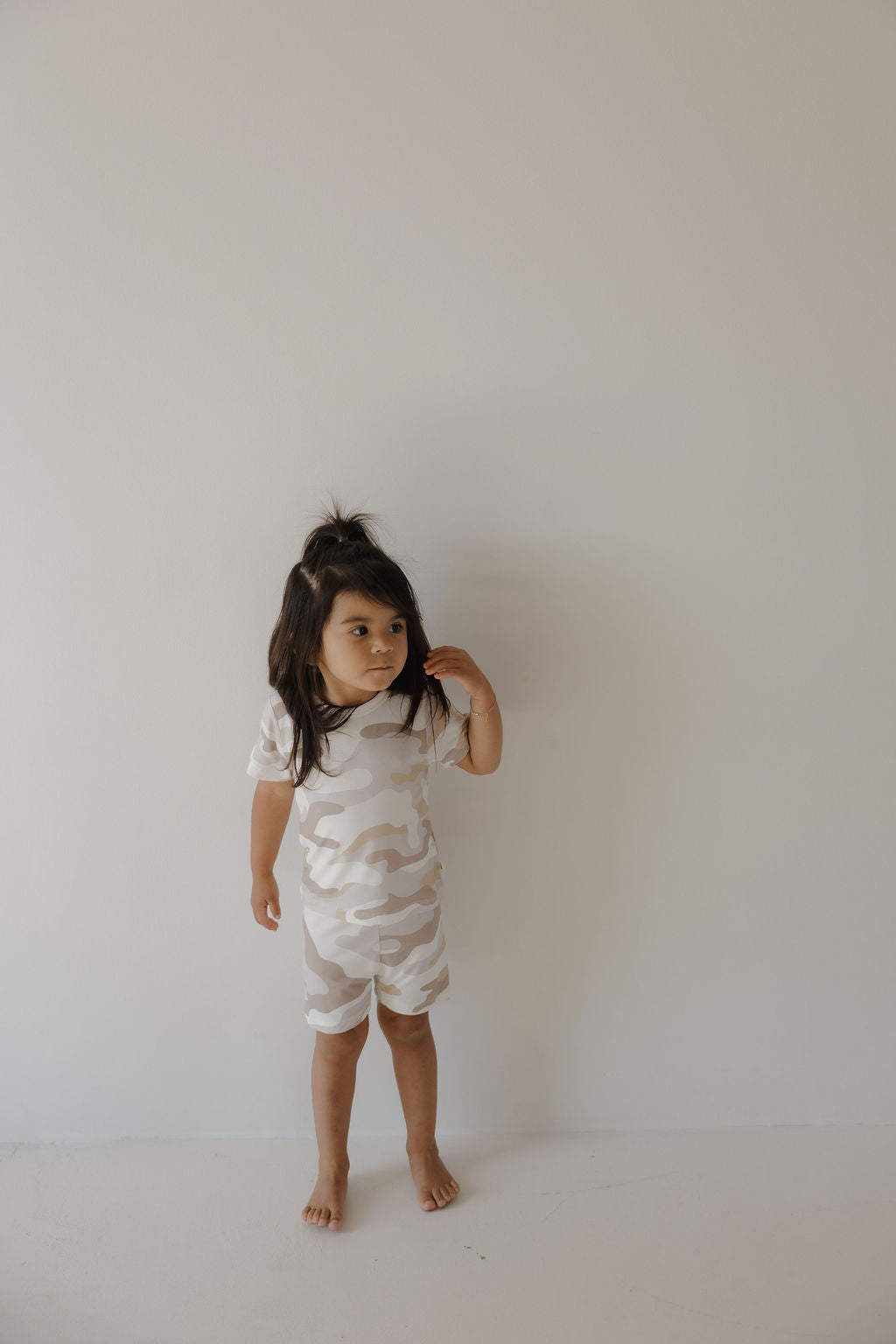 A young child with long dark hair stands barefoot against a light wall, wearing the forever french baby Bamboo Short Set | Camo 2.0—hypo-allergenic, beige and white camo top and shorts—looking to the side while touching their hair.