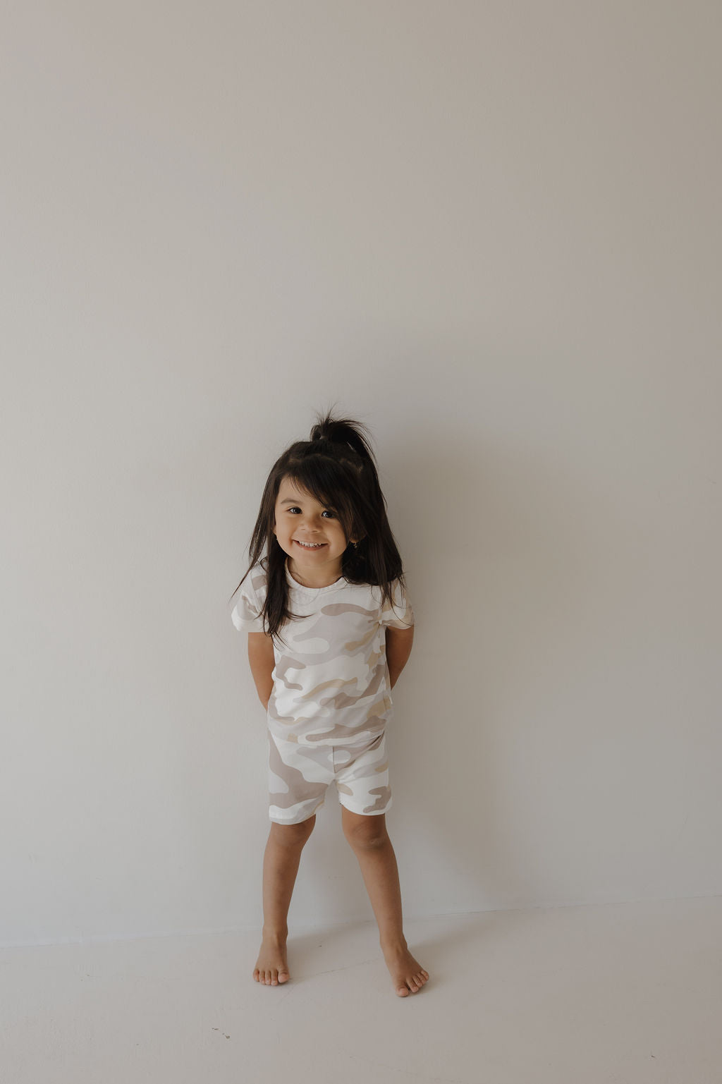 A young child with long dark hair stands barefoot against a light wall, smiling and wearing the forever french baby Bamboo Short Set | Camo 2.0, a short-sleeved, breathable camouflage outfit.