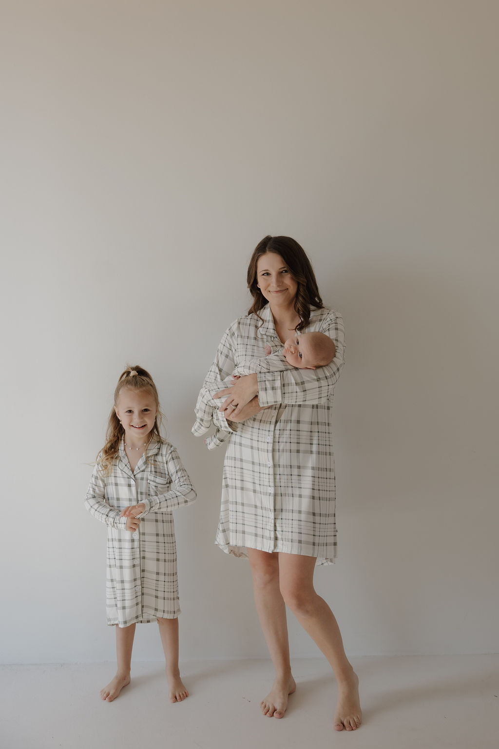A woman, a baby, and a young girl stand barefoot by a plain wall, all in matching forever french baby Toddler Bamboo Sleeping Dresses in Picnic Plaid. The woman smiles gently as she holds the baby; the girl stands with her hands clasped.
