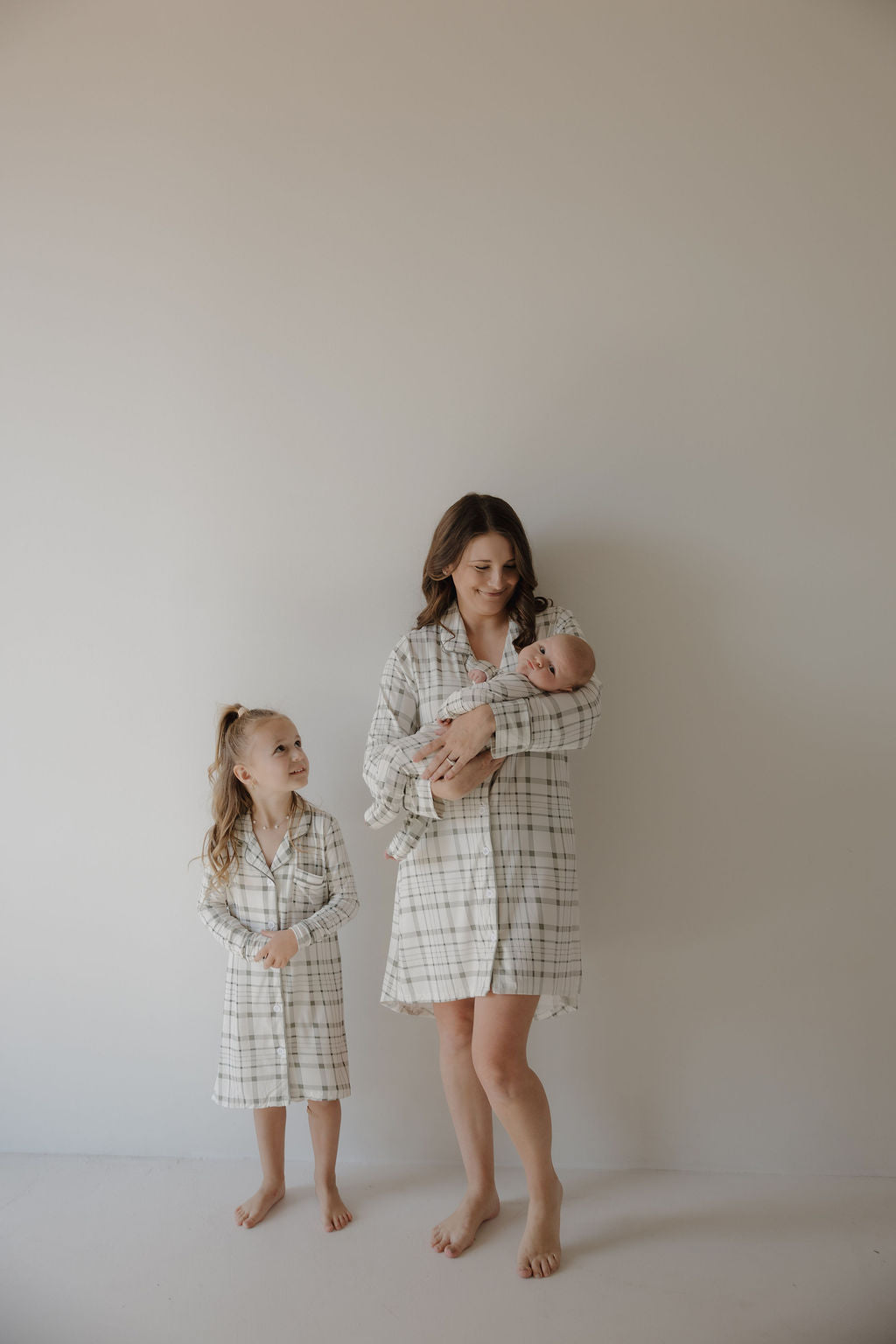 A woman with a baby stands beside a young girl, all barefoot and wearing matching forever french baby Toddler Bamboo Sleeping Dress in Picnic Plaid, made of soft bamboo fabric, against a plain light-colored wall.