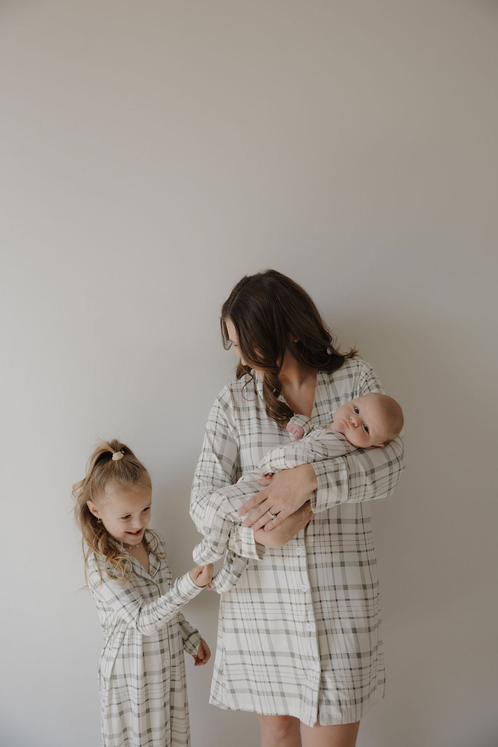 A woman holds a sleeping baby and stands next to a smiling young girl, all wearing forever french baby's Picnic Plaid bamboo clothing, including the Toddler Bamboo Sleeping Dress, against a plain light background.