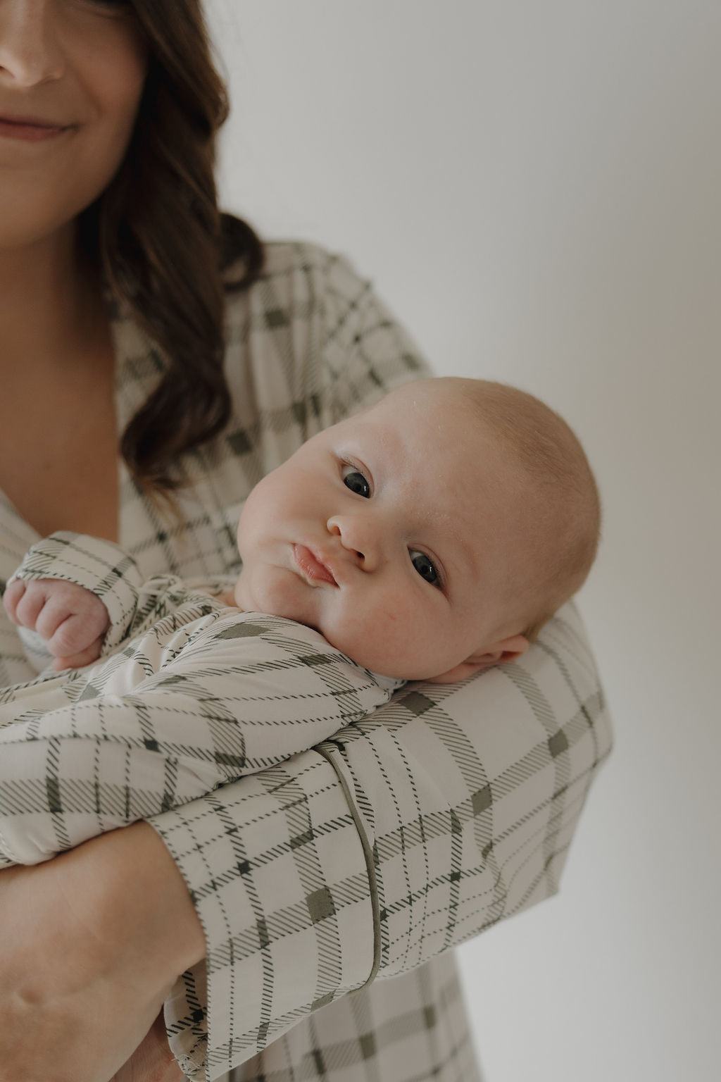 A baby in forever french baby’s plaid bamboo outfit rests calmly in an adult's arms. The adult, partly visible, wears the matching Women's Bamboo Sleeping Dress | Picnic Plaid. The soft, breathable fabric keeps the baby comfortable and serene.