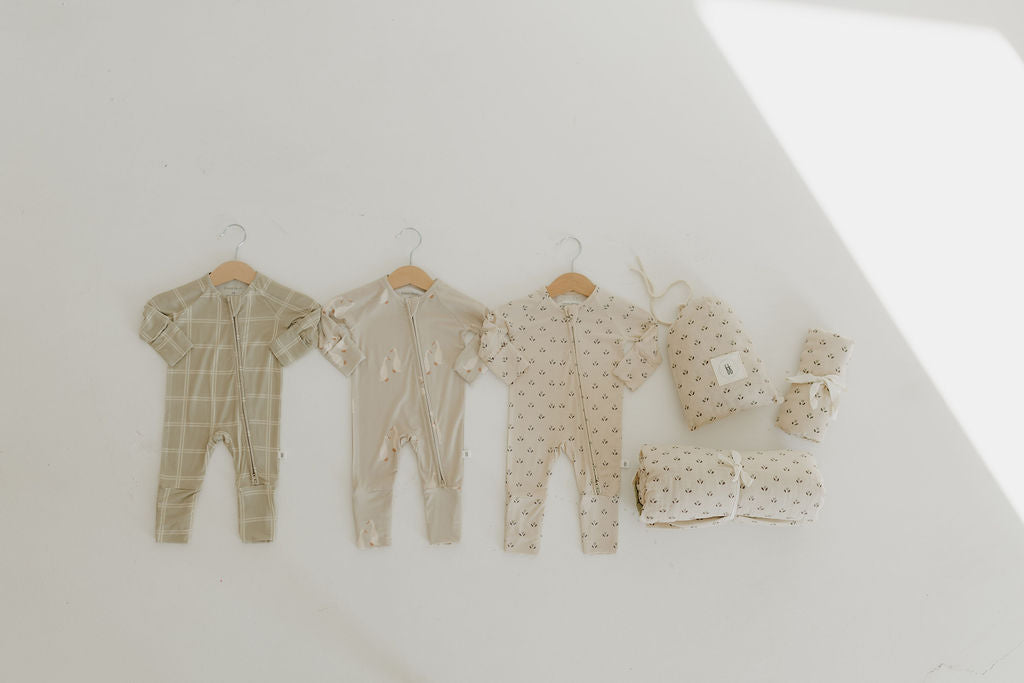 A set of three neutral baby onesies on hangers, with folded Muslin Swaddles | Flora and a patterned pillow from forever french baby, are arranged on a light floor. Soft sunlight creates gentle shadows for an inviting ambiance.