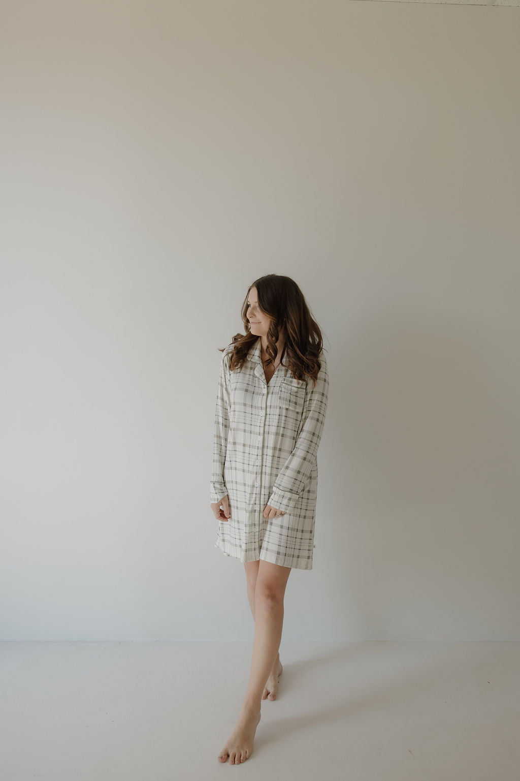 A woman with long brown hair stands barefoot on a light floor, looking left, wearing the forever french baby Women's Bamboo Sleeping Dress in Picnic Plaid.