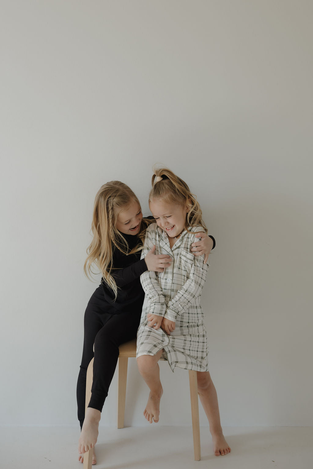 Two young girls with long blonde hair smile together; one stands barefoot in the forever french baby Toddler Bamboo Sleeping Dress in Picnic Plaid, while the other sits on a wooden stool, both set against a plain light background.