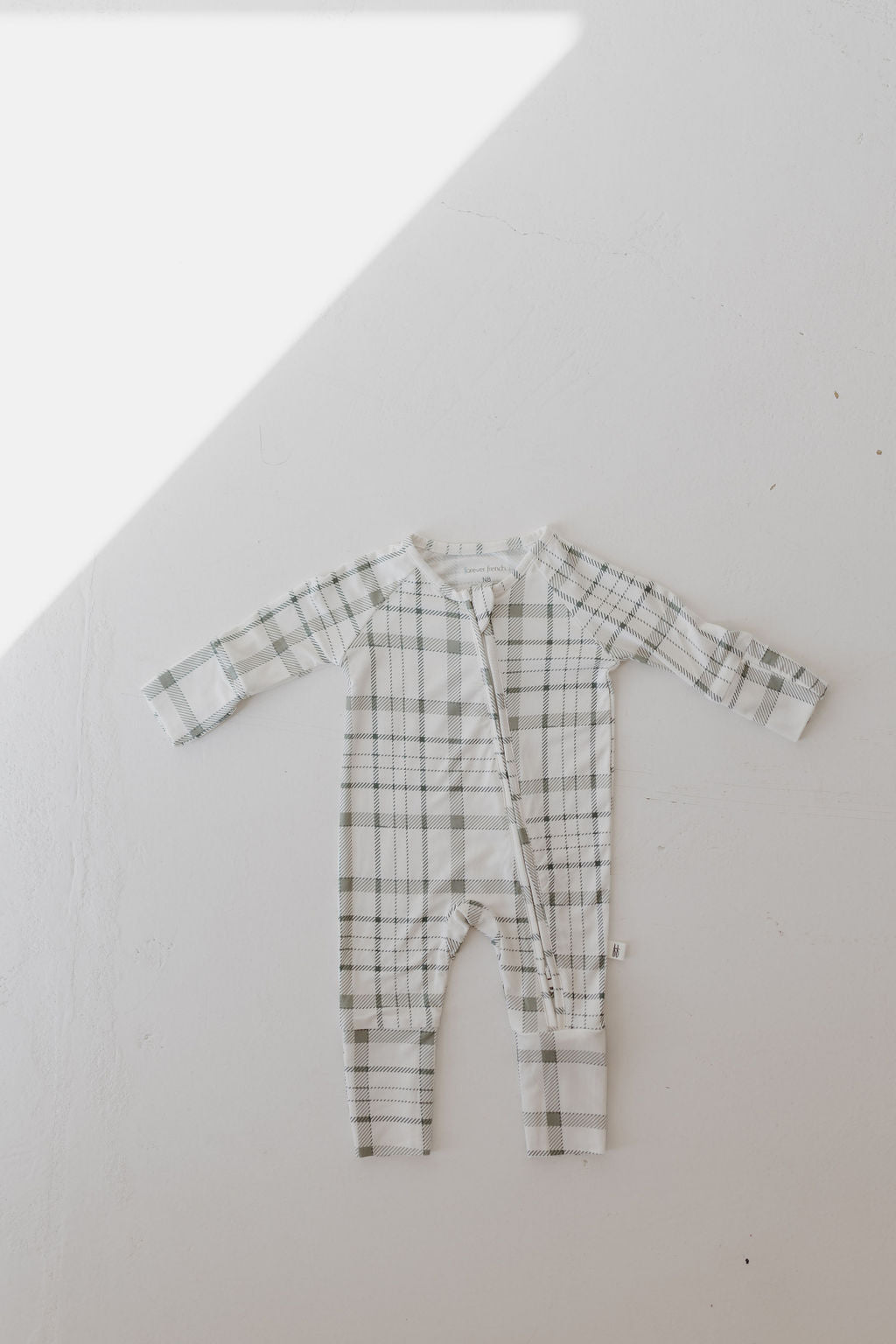 Laid out on a white surface, the Bamboo Zip Pajamas in Picnic Plaid by forever french baby feature a hypo-allergenic, green, gray, and white pattern. Sunlight casts a triangular shadow in the top left corner of the image.