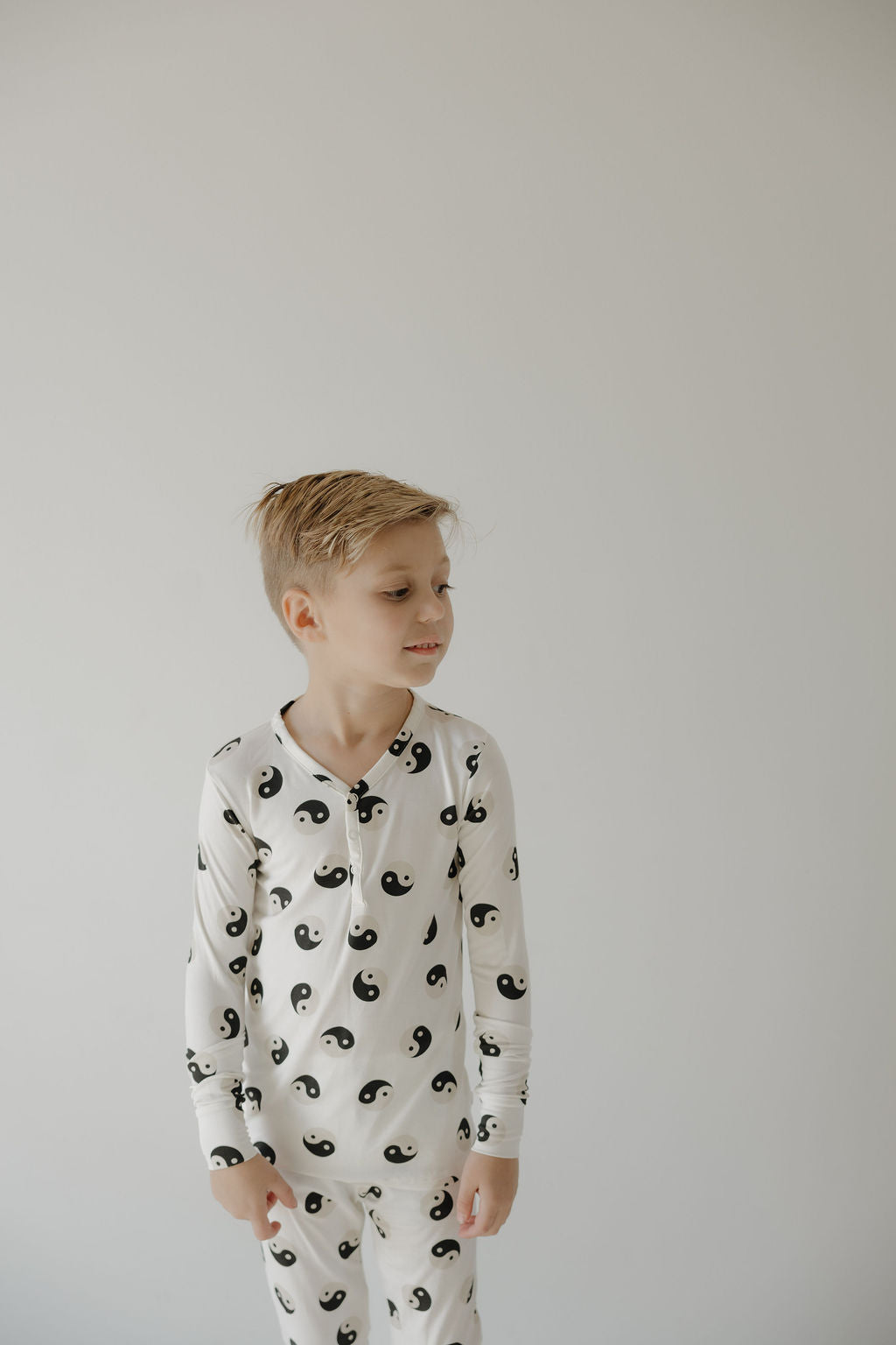 A young boy with blonde hair wears forever French baby’s Bamboo Two Piece Pajamas in Black & White Yin Yang, standing against a light background and looking left, cozy in breathable sleepwear.