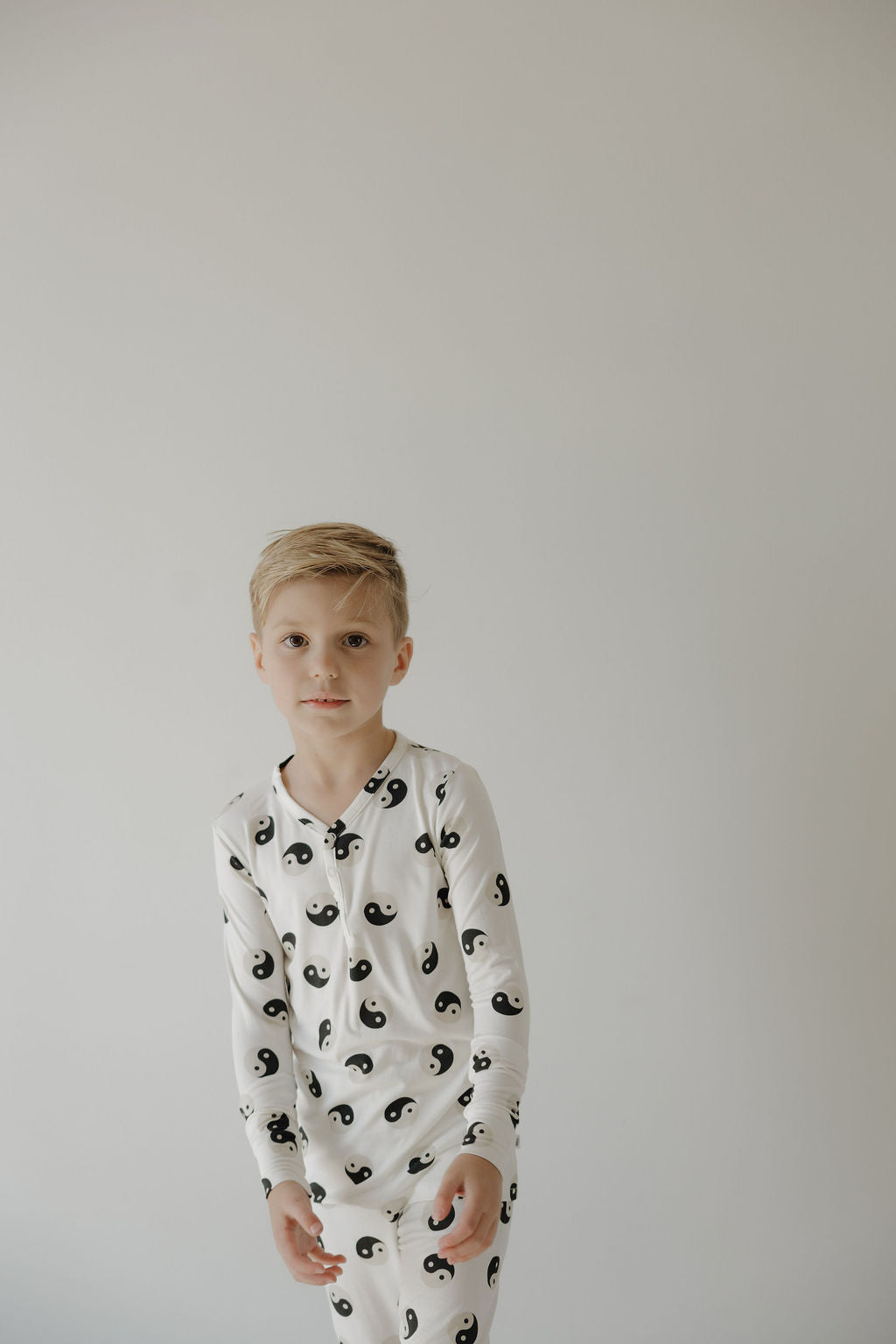 A blonde boy wears forever French baby’s Bamboo Two Piece Pajamas in Black & White Yin Yang, featuring breathable fabric and yin-yang designs, standing against a plain light background.