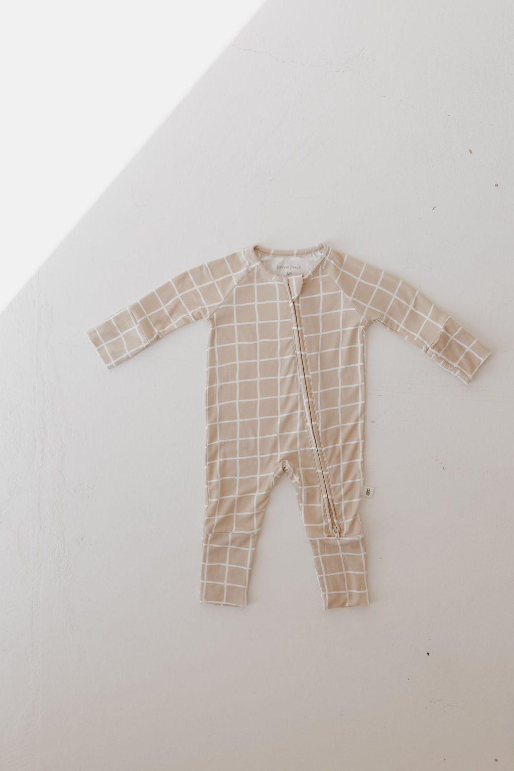 The Bamboo Zip Pajamas | Grid by forever french baby, laid flat on a light surface, features a beige and white grid pattern. Made from bamboo, this hypoallergenic sleepwear has long sleeves and legs with a front zipper, subtly highlighted by soft diagonal lighting.