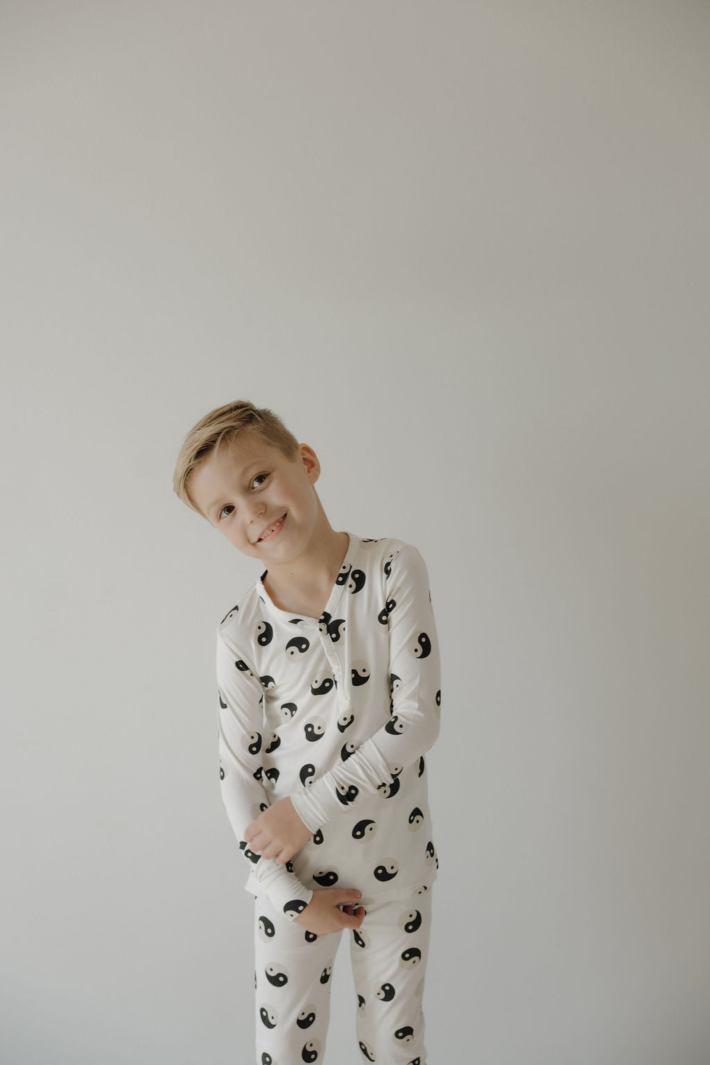 A young blond boy smiles in forever French baby's Bamboo Two Piece Pajamas | Black & White Yin Yang, made with breathable, hypo-allergenic fabric for comfort against a plain light background.