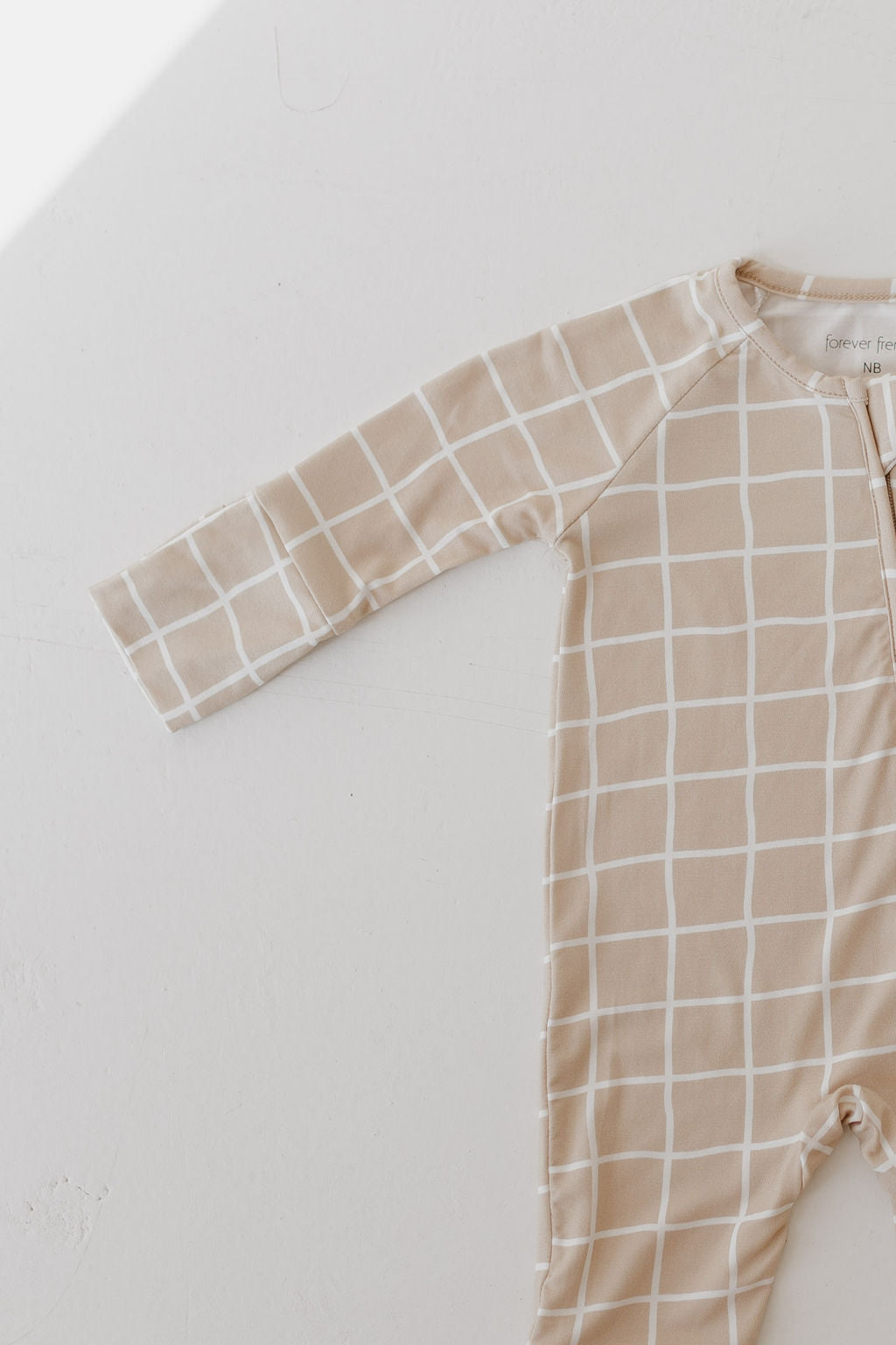 The Bamboo Zip Pajamas | Grid from forever french baby features a beige design with a white grid pattern. Photographed with natural lighting on a white background, this hypoallergenic bamboo sleepwear shows its breathable texture and design, revealing one sleeve and part of the body.