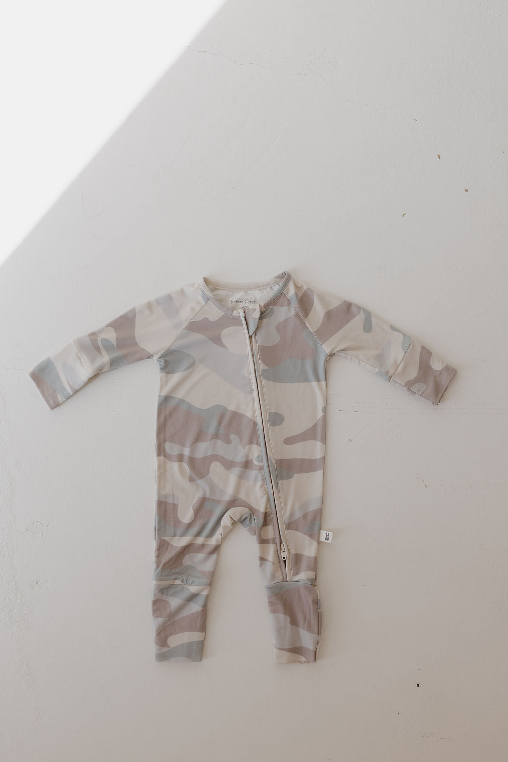 The Bamboo Zip Pajamas in the Camo pattern by forever french baby is a breathable sleepwear onesie featuring long sleeves and a diagonal zipper from neckline to left leg. It lies flat on a white surface, with a shadow across the top left corner.