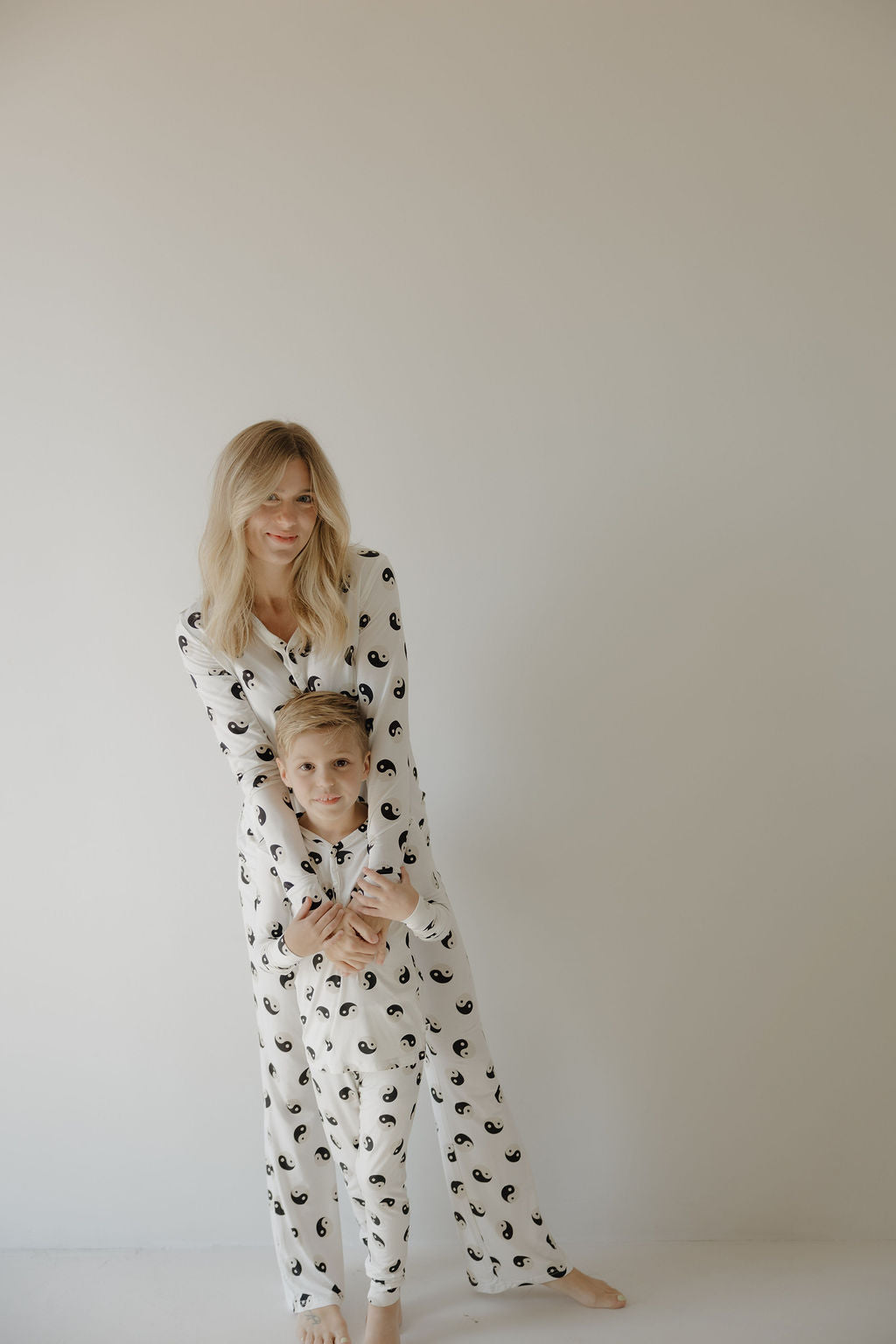 A woman and young boy wear matching forever French baby Black & White Yin Yang bamboo pajamas, standing together in a softly lit room as she gently holds him with a warm, affectionate smile.