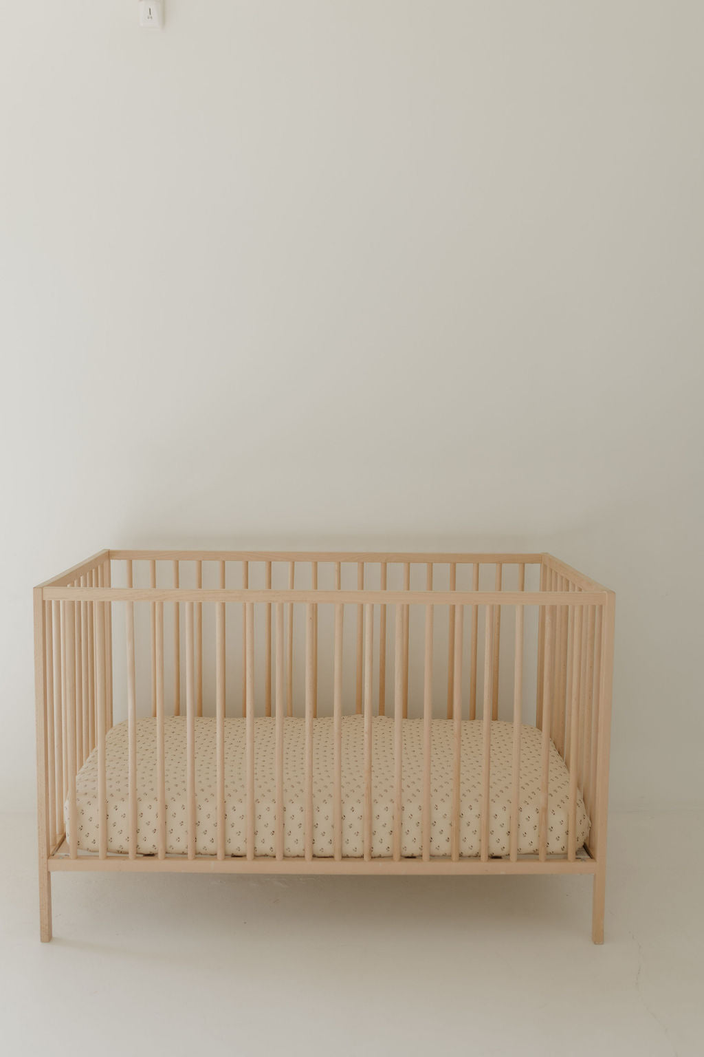 A minimalist nursery showcases a light wooden crib with vertical slats against a plain white wall. Inside, a mattress is draped with the Muslin Crib Sheet | Flora by forever french baby, featuring beige polka dots. The room is softly lit, enhancing its simplicity and cleanliness.