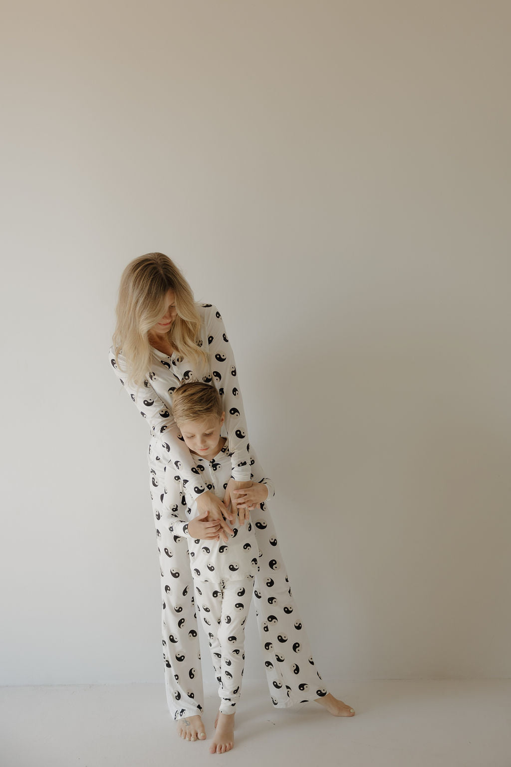 A woman and a young boy wear matching forever French baby Women's Bamboo Pajamas in the Black & White Yin Yang design as they stand barefoot against a plain light wall, with the woman gently hugging the boy from behind.