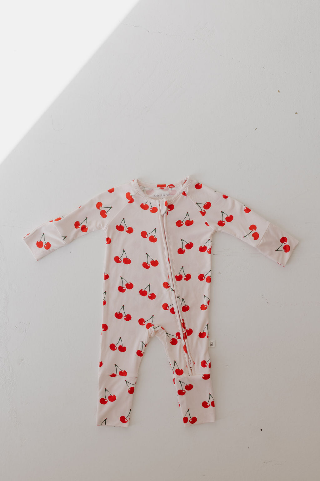 The Bamboo Zip Pajamas | Cherry Orchard by forever french baby is laid flat on a white surface. Made from hypo-allergenic material, it showcases a cheerful red cherries pattern with a front zipper. Light casts a diagonal shadow across the image, accentuating its breathable fabric.