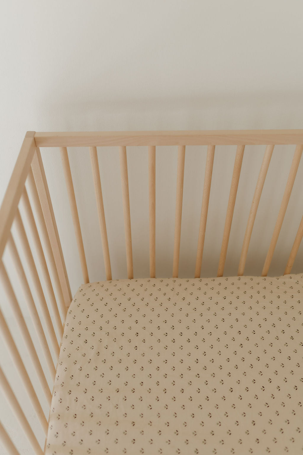 A wooden crib with light-colored bars features the Muslin Crib Sheet | Flora by forever french baby, showcasing a subtle beige pattern. It's set against a plain white wall.