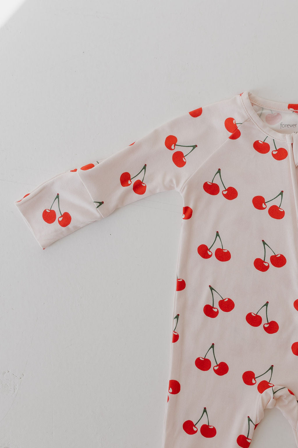 The forever french baby Bamboo Zip Pajamas in the Cherry Orchard pattern are crafted from hypo-allergenic fabric. This breathable, long-sleeve onesie features a charming design of red cherries with green stems on a white background.