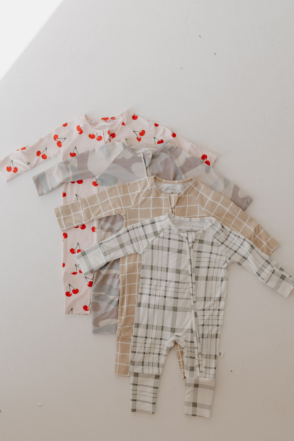 Four folded baby onesies from forever french baby are on a white surface: the top one is made of breathable sleepwear with red cherries, the second has a neutral camouflage pattern, the third is a Bamboo Zip Pajamas with a beige grid pattern, and the bottom features green and white plaid.