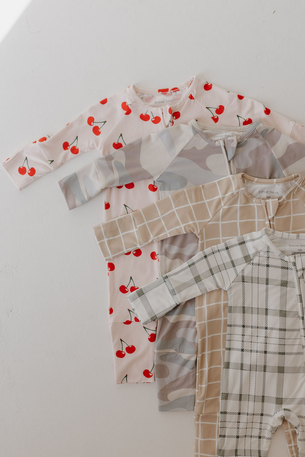 A set of four long-sleeve baby onesies from forever french baby is displayed. The hypoallergenic bamboo zip pajamas feature designs with red cherries on white, gray camouflage, beige with white grid lines, and green plaid with light beige details—ideal for delicate skin.