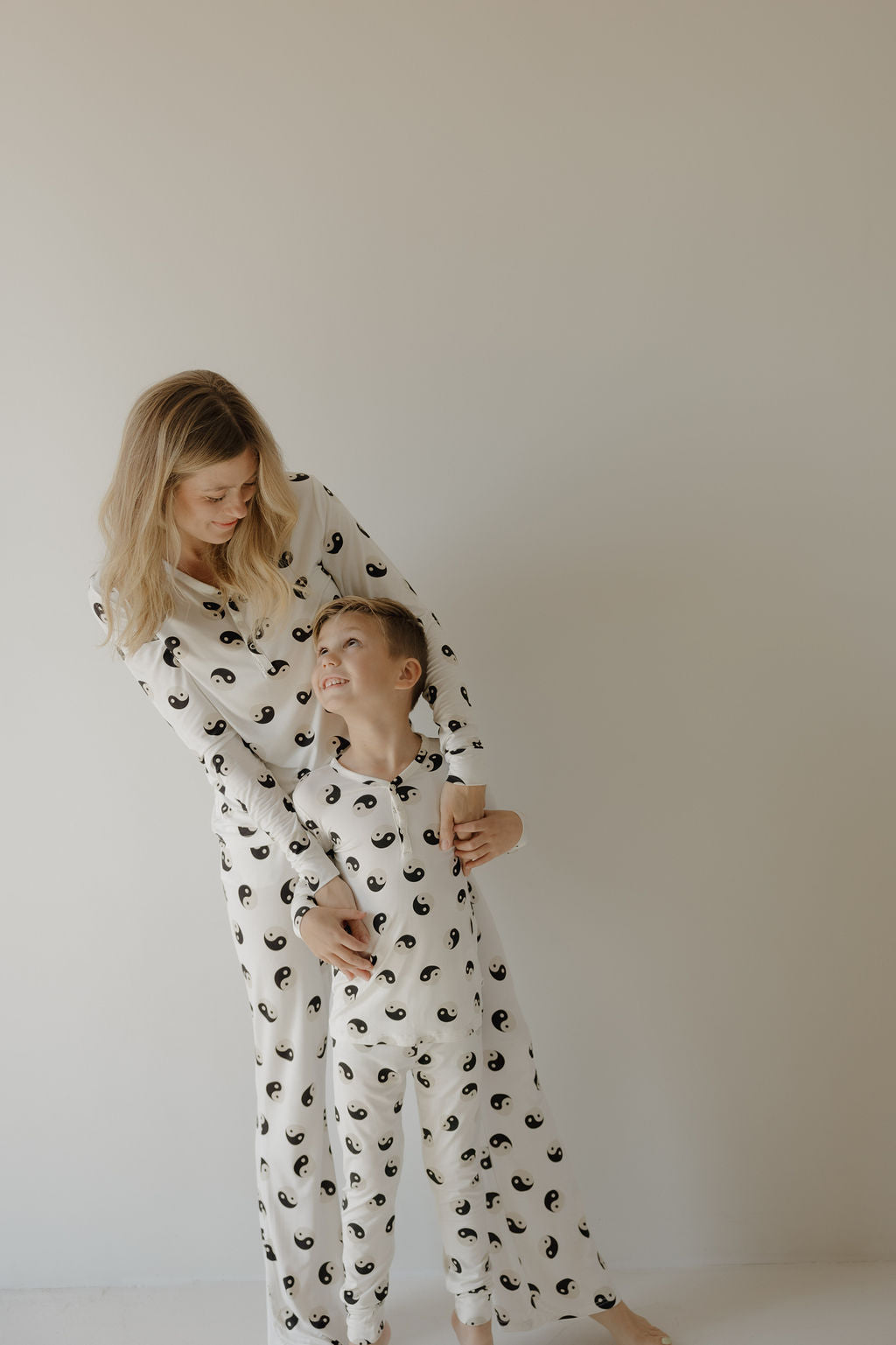 A woman and a young boy smile at each other in matching forever French baby Women's Bamboo Pajama sets, displaying the black & white Yin Yang design, against a plain light background.