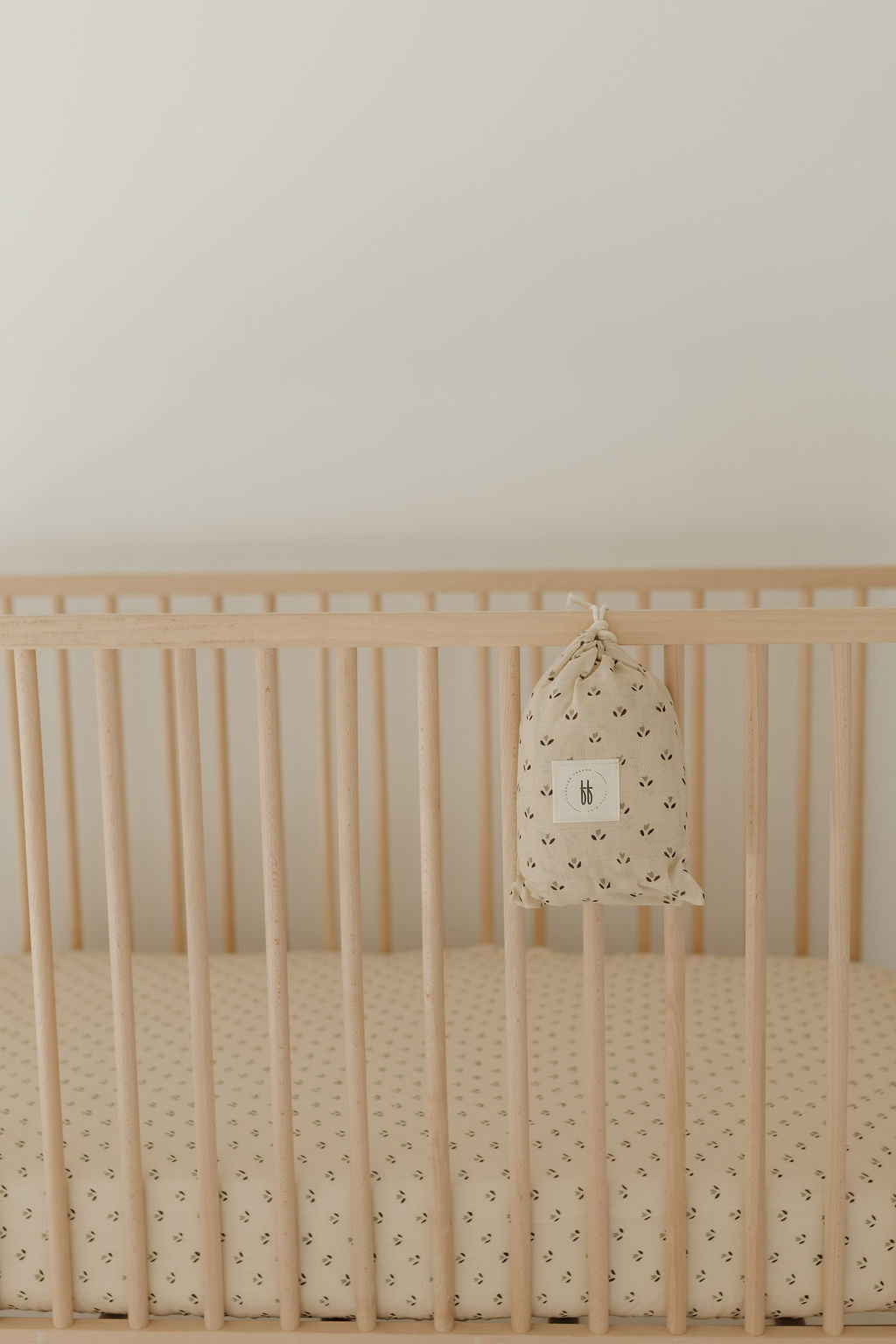 A wooden baby crib with the "Muslin Crib Sheet | Flora" by forever french baby, showcasing a light tan mattress and subtle floral design. A matching fabric bag with a small design is tied to the side rail, set against a plain, light-colored wall.