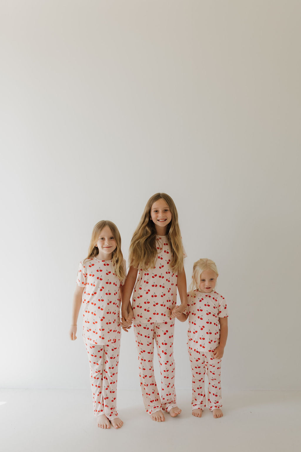 Three smiling children stand against a plain white background in matching Toddler Short Sleeve Bamboo Lounge Sets from forever french baby, featuring a cherry orchard print. The tallest in the middle holds hands with the others on either side.