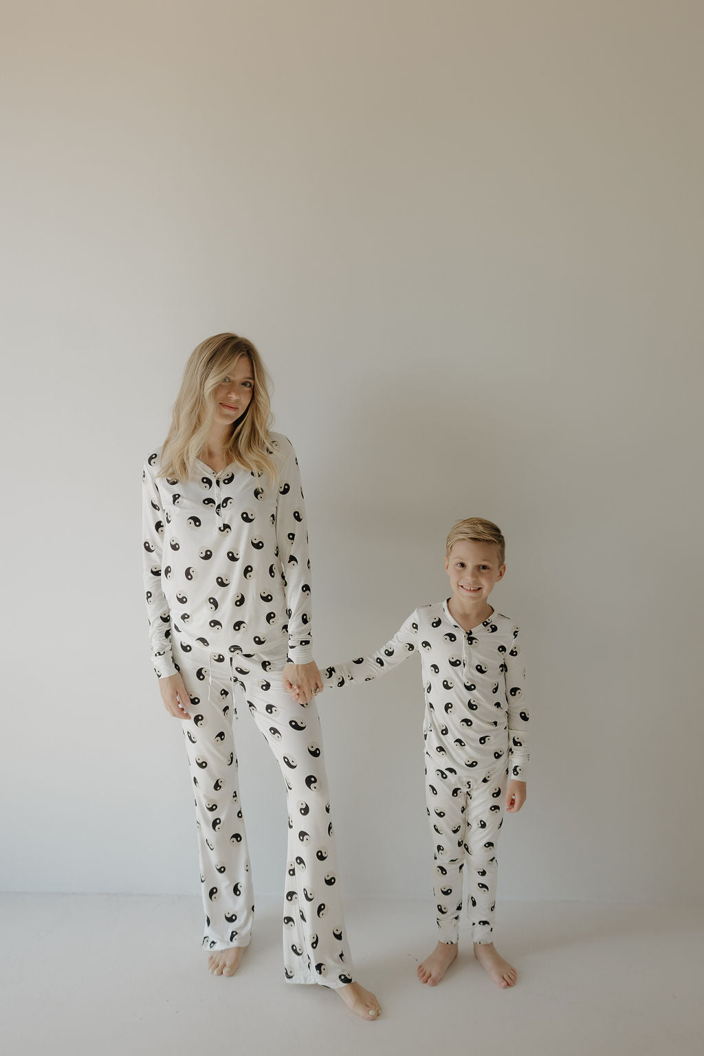 A woman and young boy, both barefoot, hold hands against a light background, wearing matching forever French baby Women's Bamboo Pajama sets in a Black & White Yin Yang abstract design.