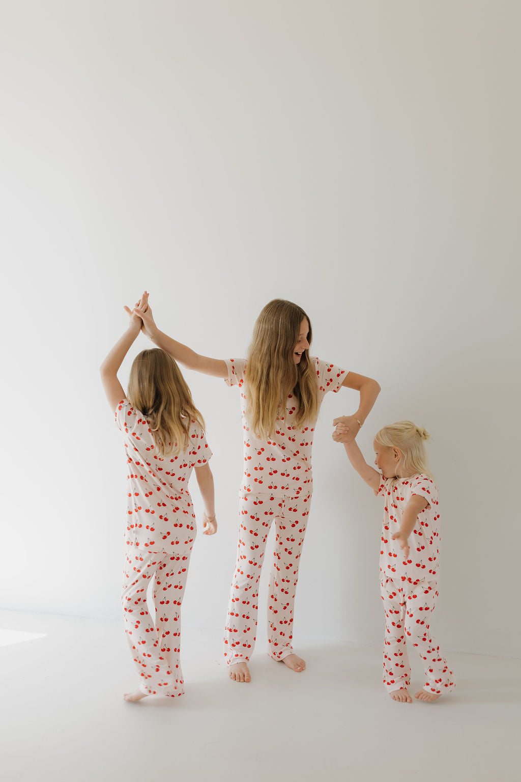 Three children wear matching forever french baby Toddler Short Sleeve Bamboo Lounge Sets in Cherry Orchard, standing barefoot against a white background. The two older kids joyfully play with the youngest, creating a cheerful and playful atmosphere.