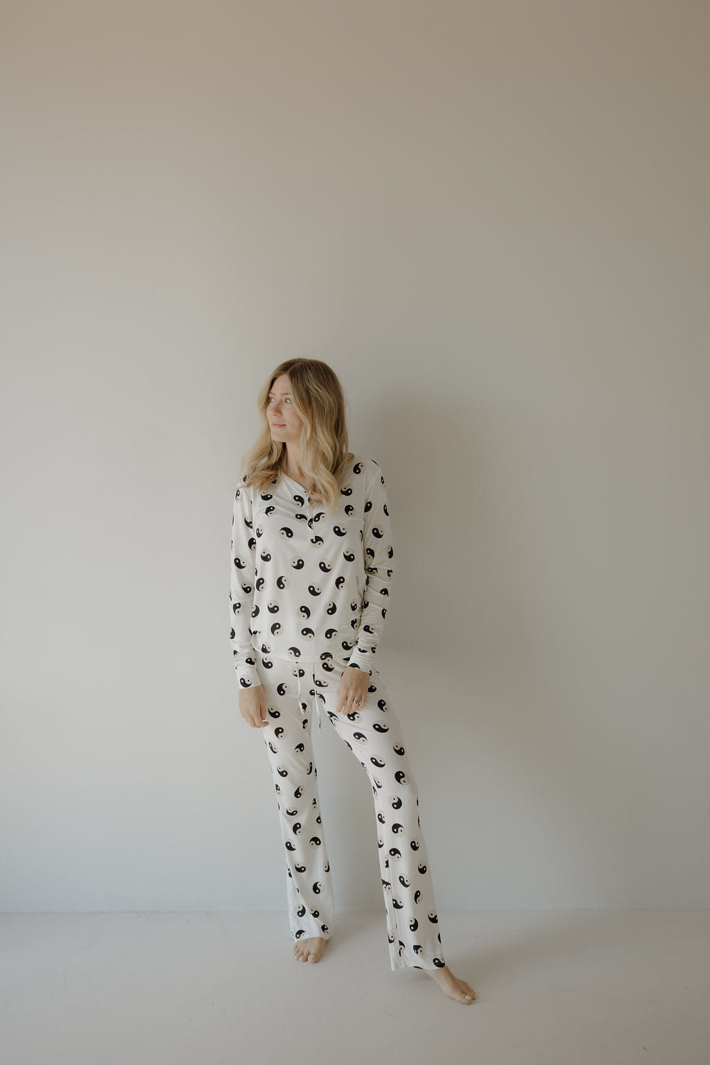 A woman with long, wavy blonde hair stands barefoot against a light wall, wearing forever French baby's Women's Bamboo Pajama in Black & White Yin Yang—a breastfeeding-friendly set. She looks left, relaxed, hands at her sides.