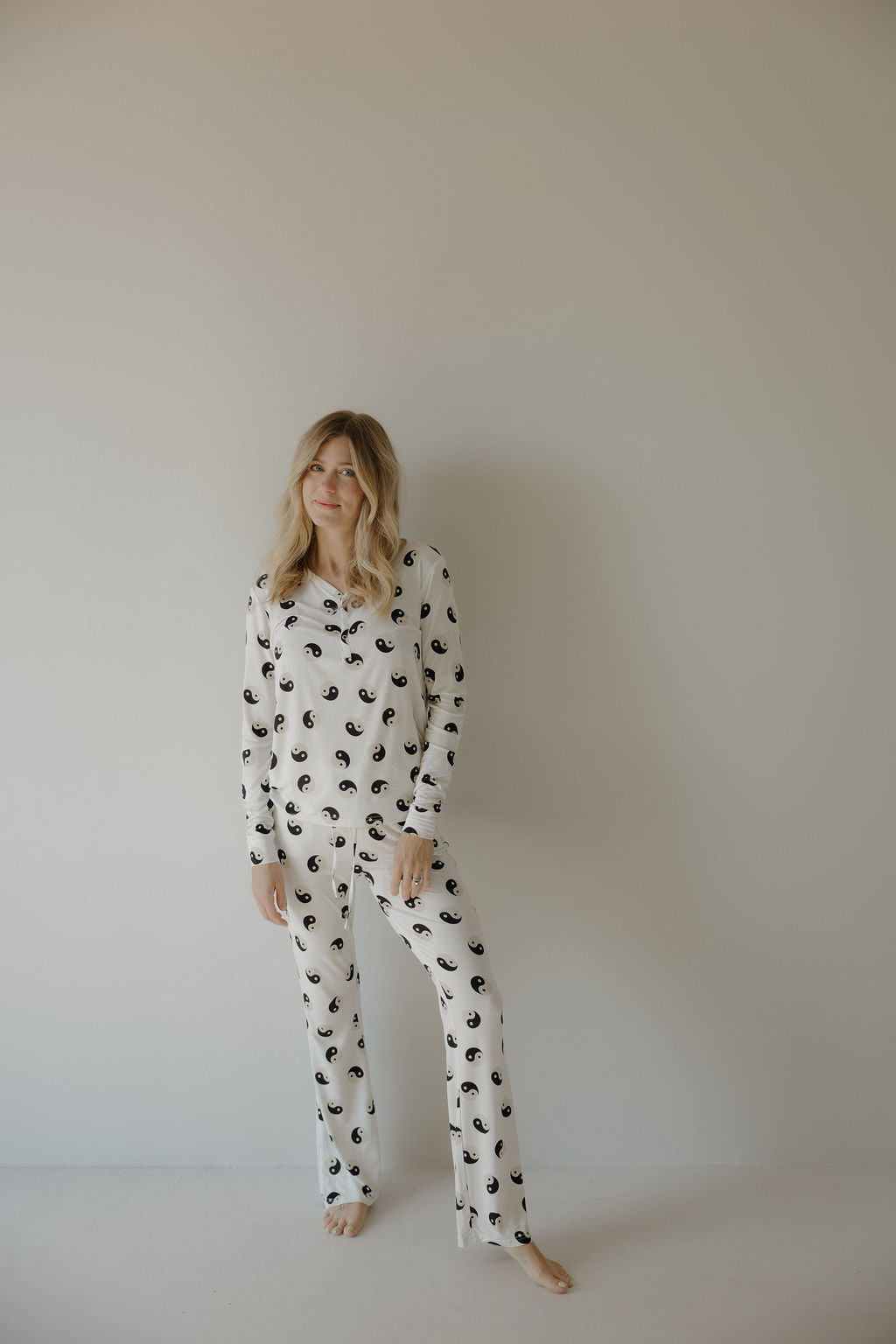 A woman with long blonde hair stands barefoot against a plain light wall, wearing forever French baby's Women's Bamboo Pajama in Black & White Yin Yang. She has a relaxed posture and gentle expression, showcasing the breastfeeding-friendly design.