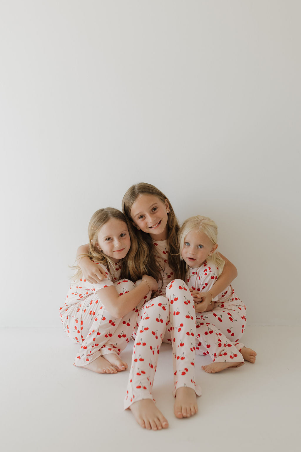 Three young girls sit on the floor against a plain white background, wearing matching Toddler Short Sleeve Bamboo Lounge Sets in Cherry Orchard from forever french baby. The breathable fabric is adorned with red cherry patterns. They are close together, smiling as the older ones hug the younger child.