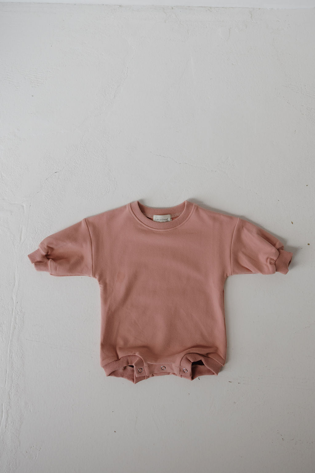 The forever french baby Sweatshirt Romper | Rose, a long-sleeved dusty pink baby romper with bottom snap buttons, is laid flat on a textured white surface.