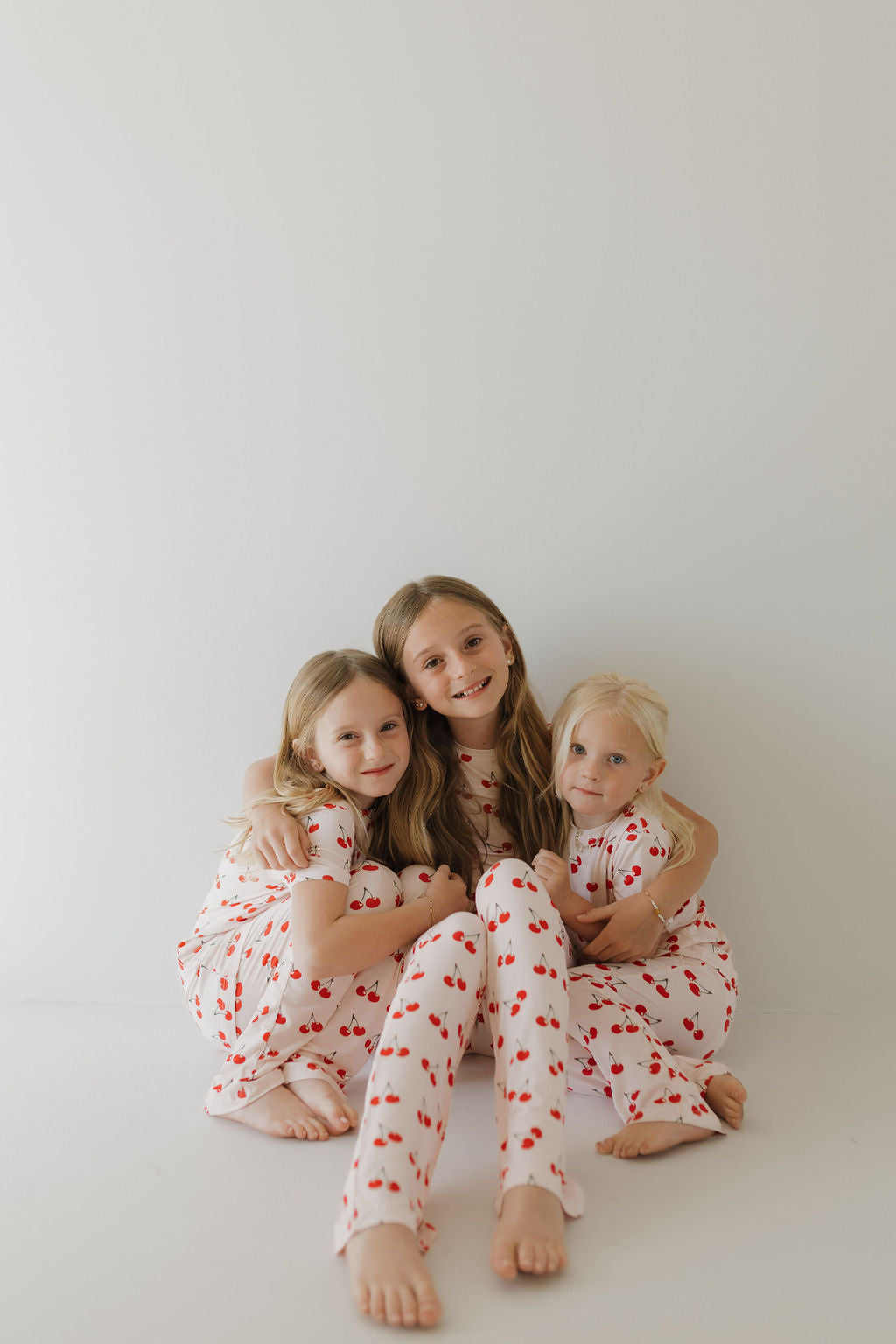 Three children sit against a plain background, all wearing matching Toddler Short Sleeve Bamboo Lounge Sets in Cherry Orchard design by forever french baby. Made from breathable fabric, they're smiling with arms around each other, appearing happy and relaxed.
